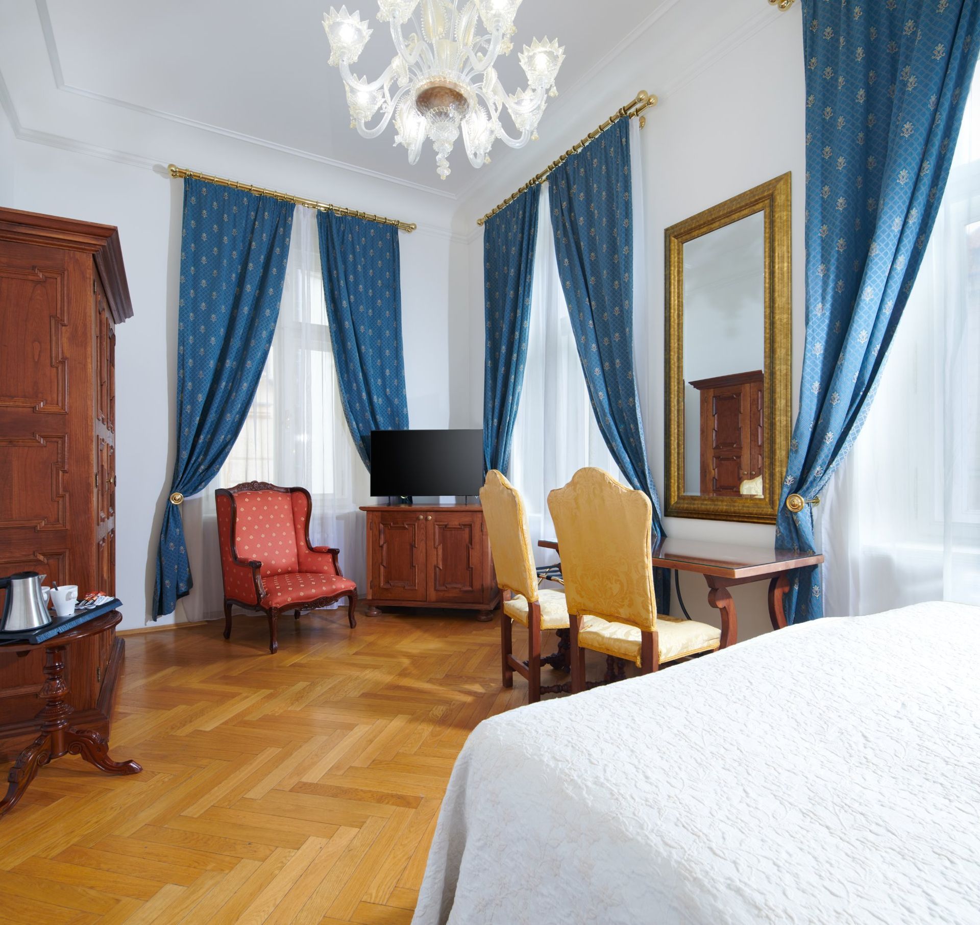 Elegant hotel room with hardwood floors, chandelier, blue curtains, wooden furniture, and a red armchair.