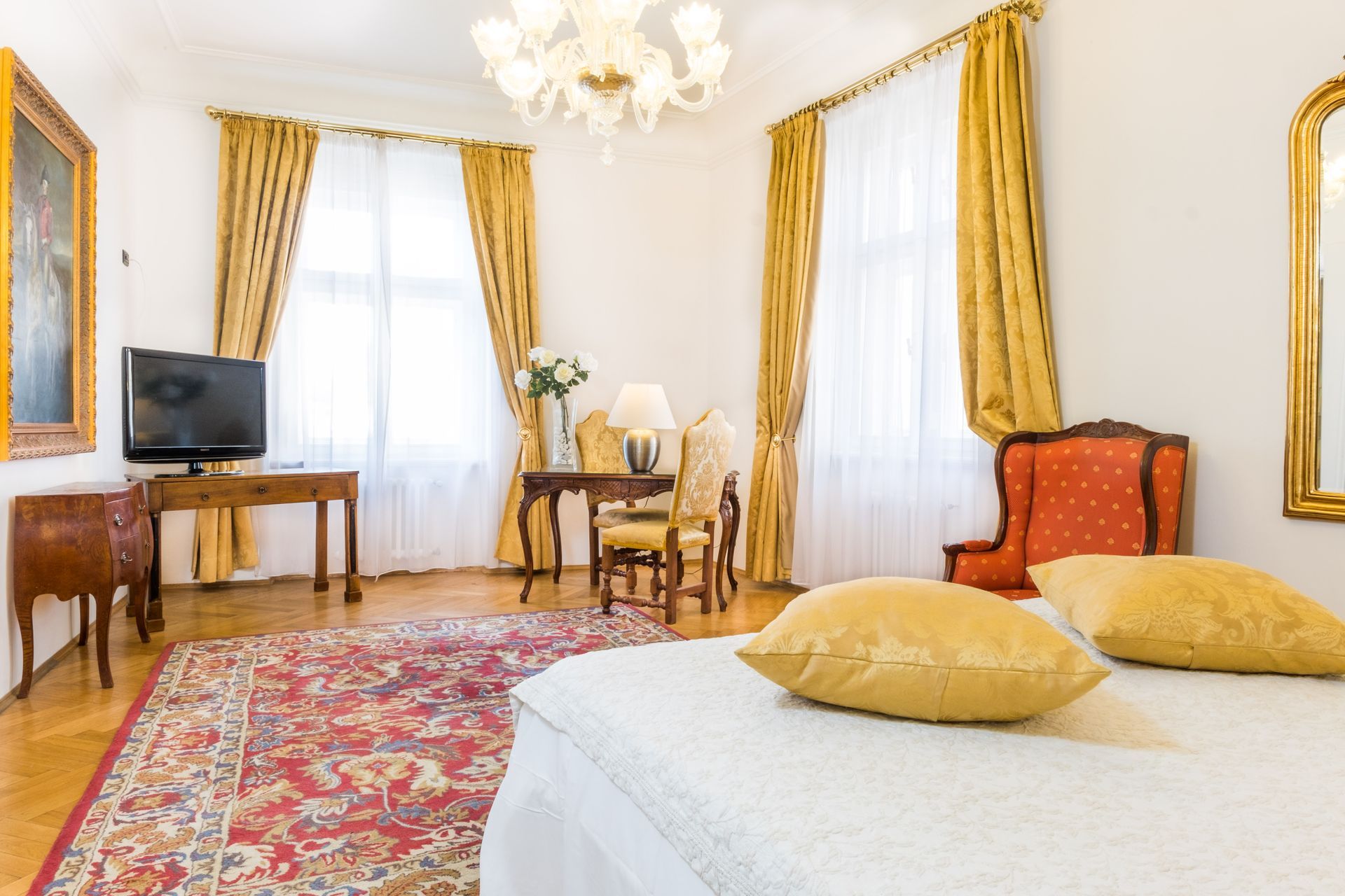 Elegant hotel room with bed, table, red rug, and gold curtains.