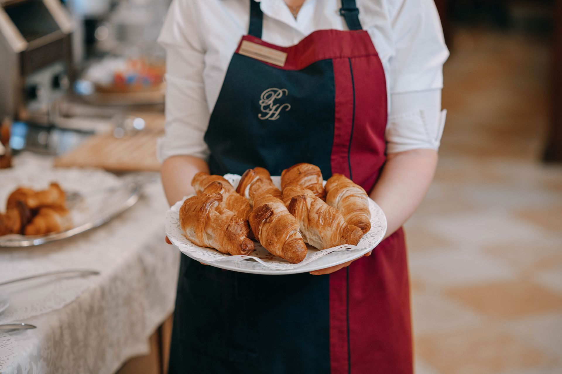 Person in apron holding plate of croissants. Table with more pastries in background.