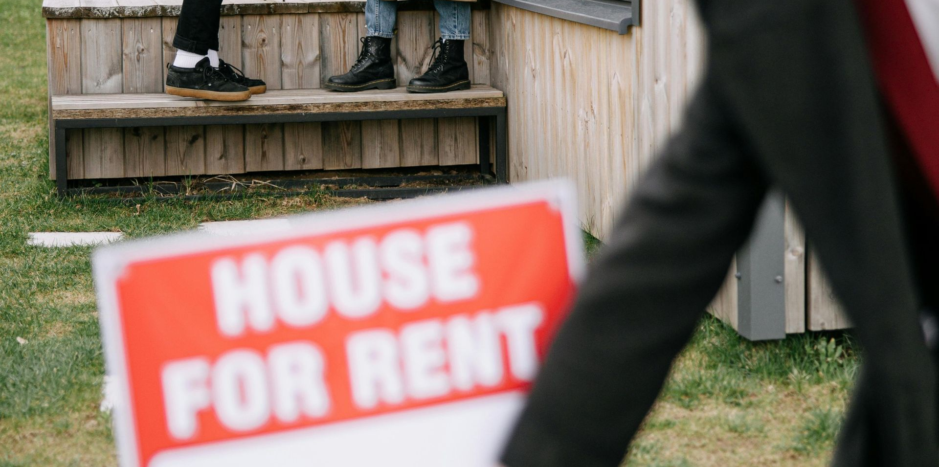 Two people talking on a porch beside a house for rent sign.