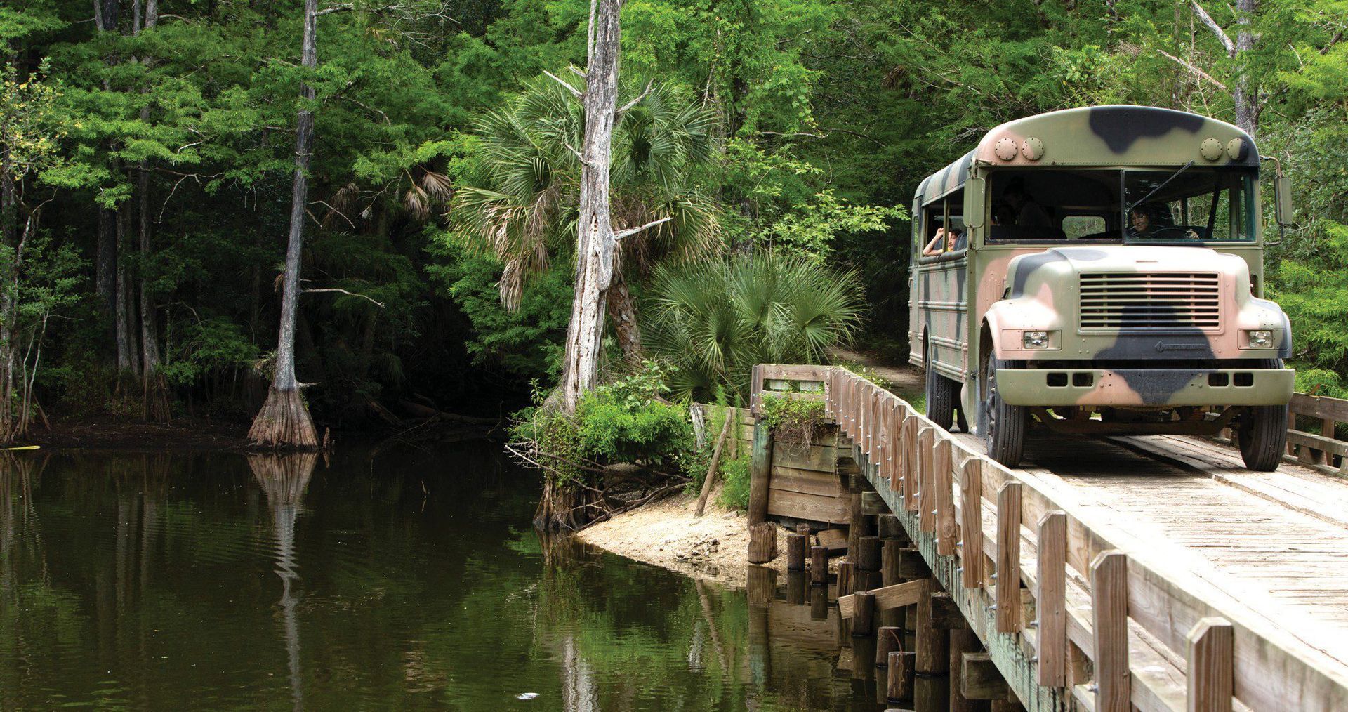 Camouflage bus driving across a wooden bridge over a swamp. Trees and water in the background.