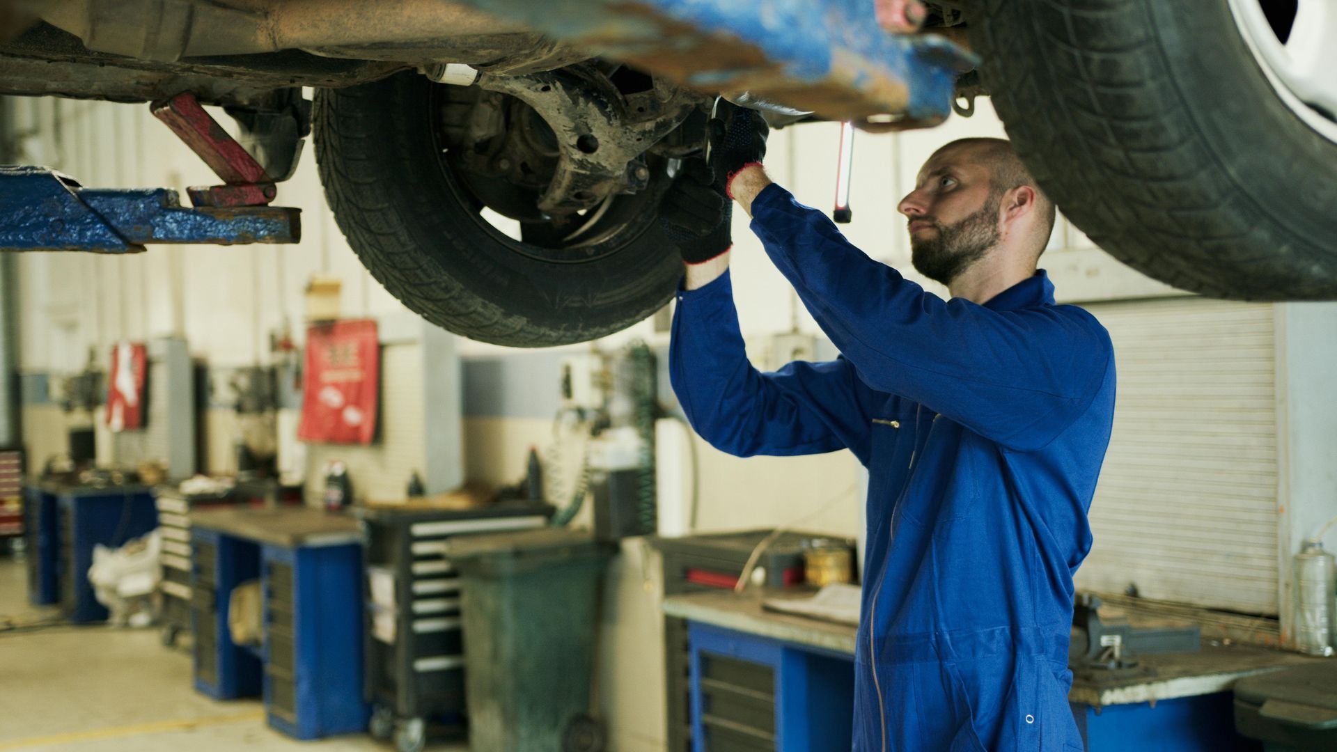 Mechanic in blue jumpsuit working under a car raised on a lift in a garage.