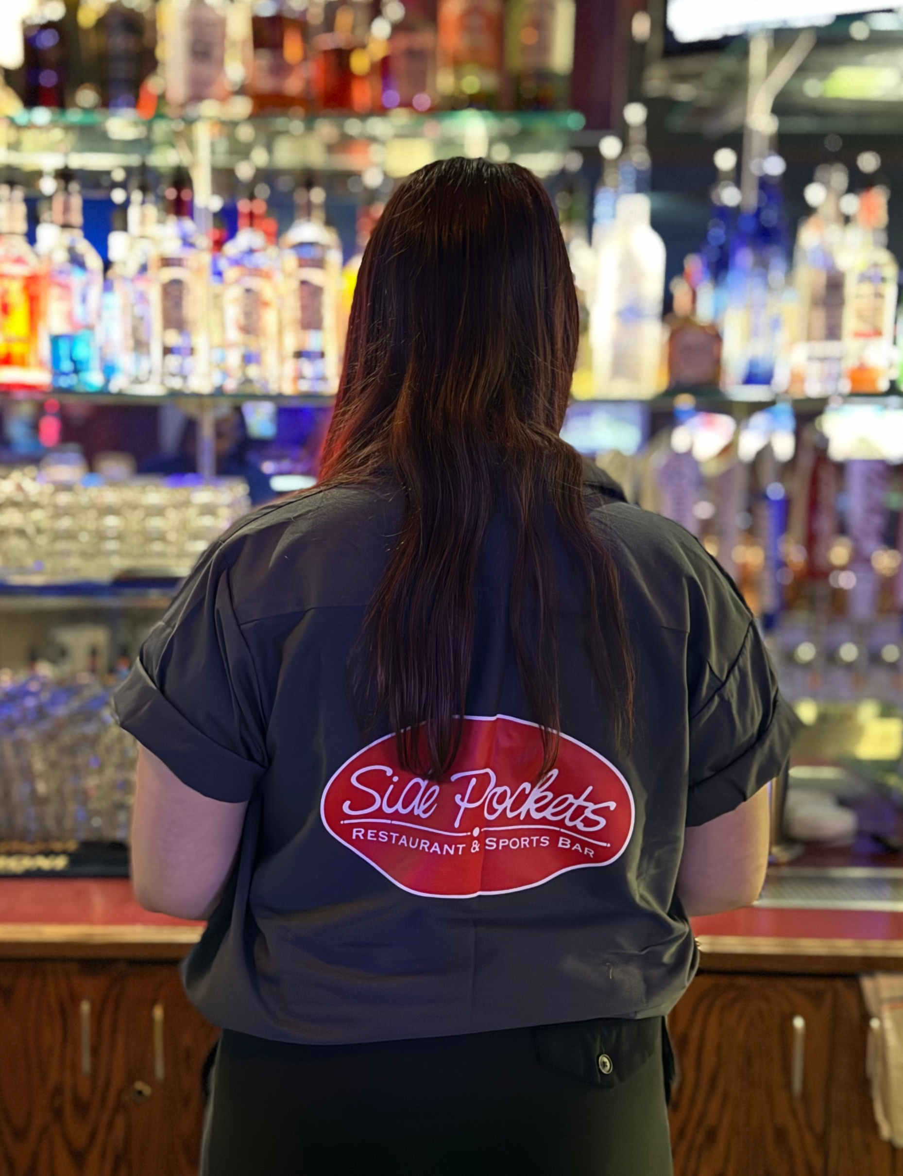 A woman wearing a shirt that says side pockets is standing in front of a bar at Side Pockets Wichita
