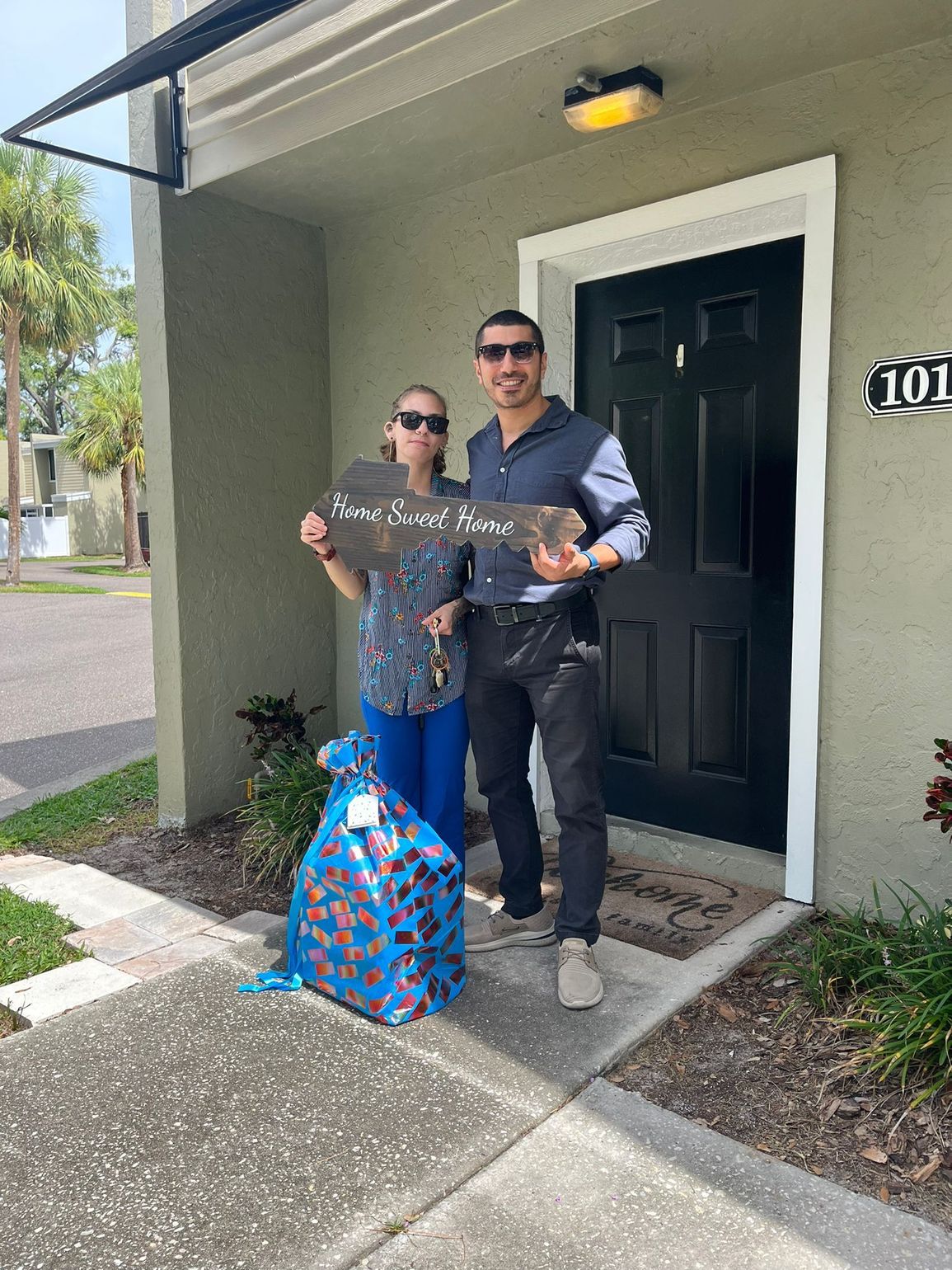 Two people holding a “thank you” sign and a blue gift bag at a front door.