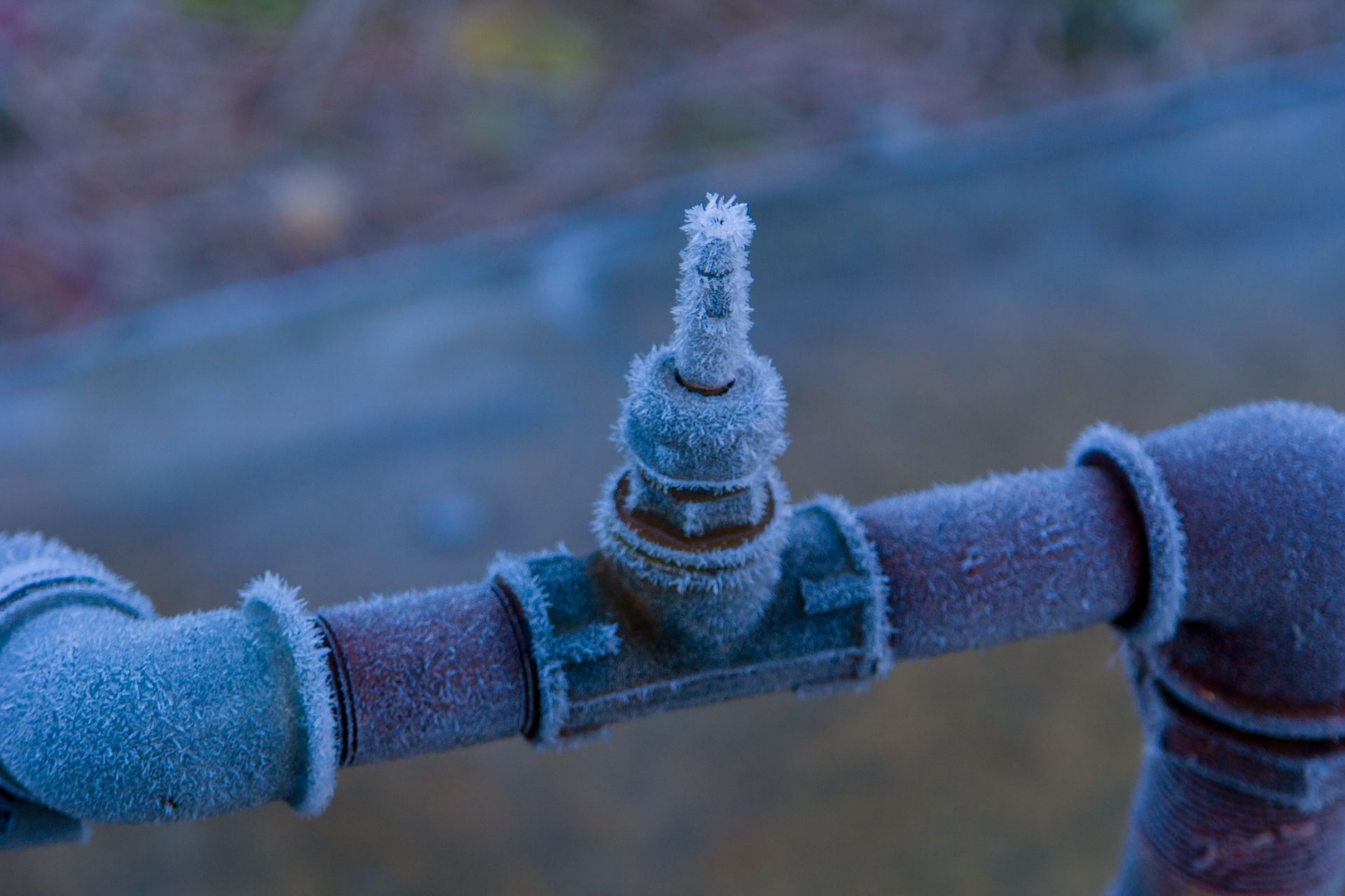 Frost-covered pipe valve outdoors, coated in ice crystals, with a blurred blue background.
