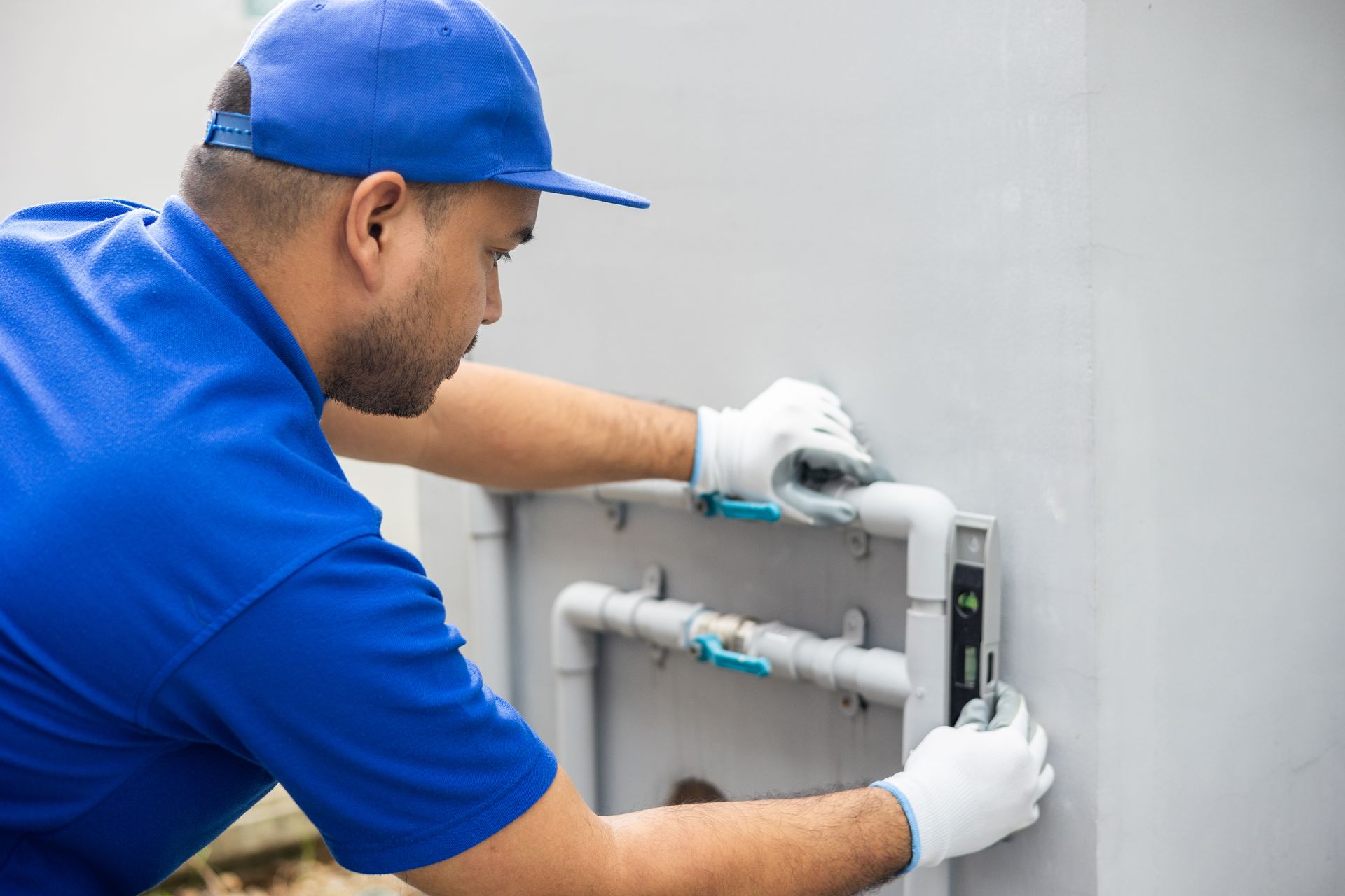 Plumber in blue shirt and cap, white gloves, working on pipes outdoors.