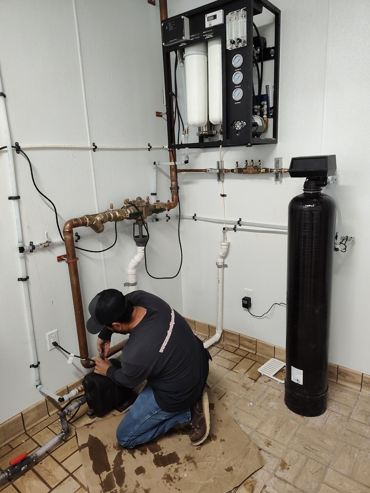 A technician works on plumbing pipes and a water filtration system mounted on a white wall in a utility room.