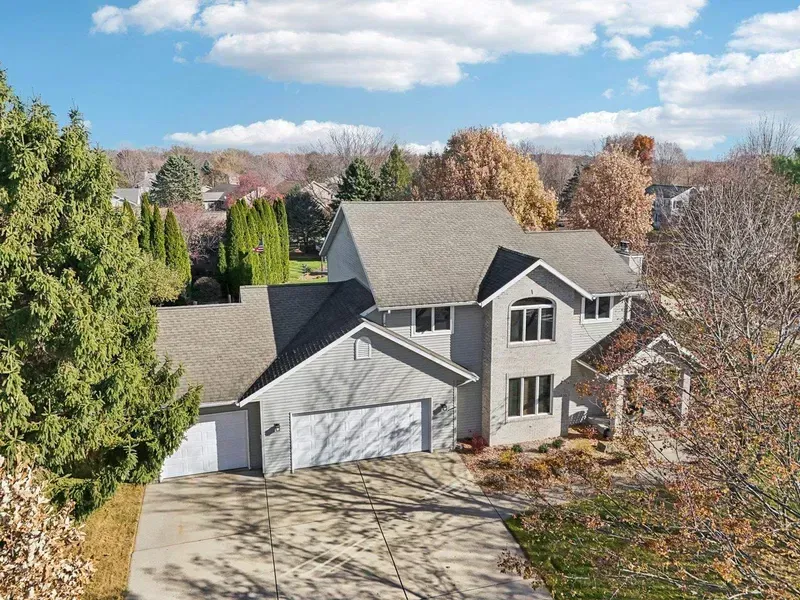 Gray two-story house with a two-car garage, driveway, and autumn trees under a blue sky.