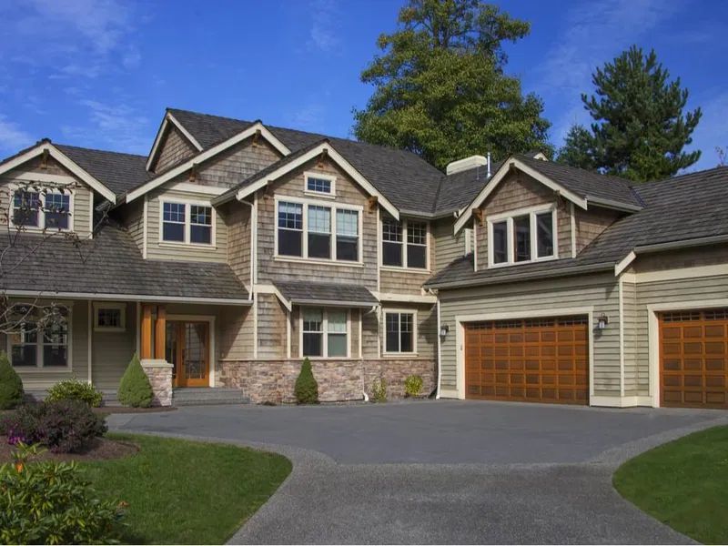 Large house with multiple gabled roofs, garage doors, and a winding driveway against a blue sky.