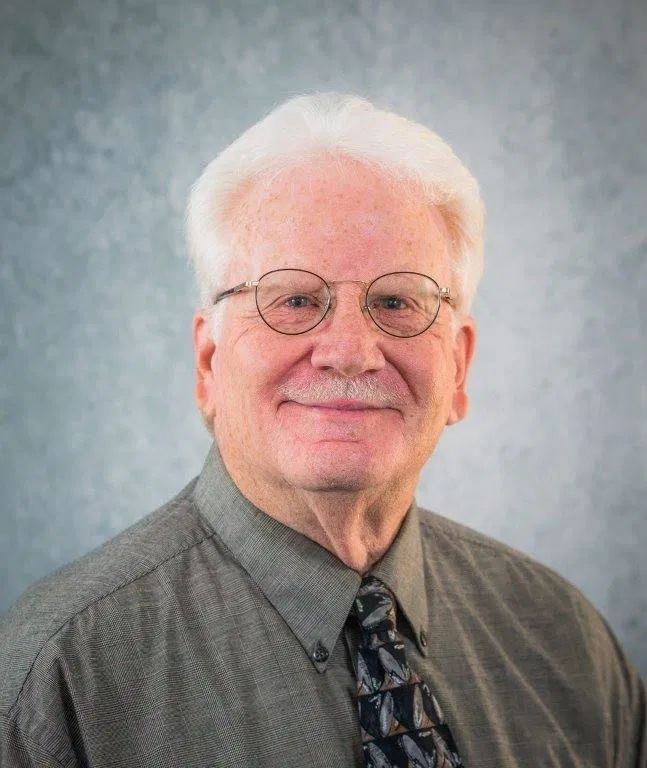 Man wearing glasses, gray shirt, and patterned tie smiling.