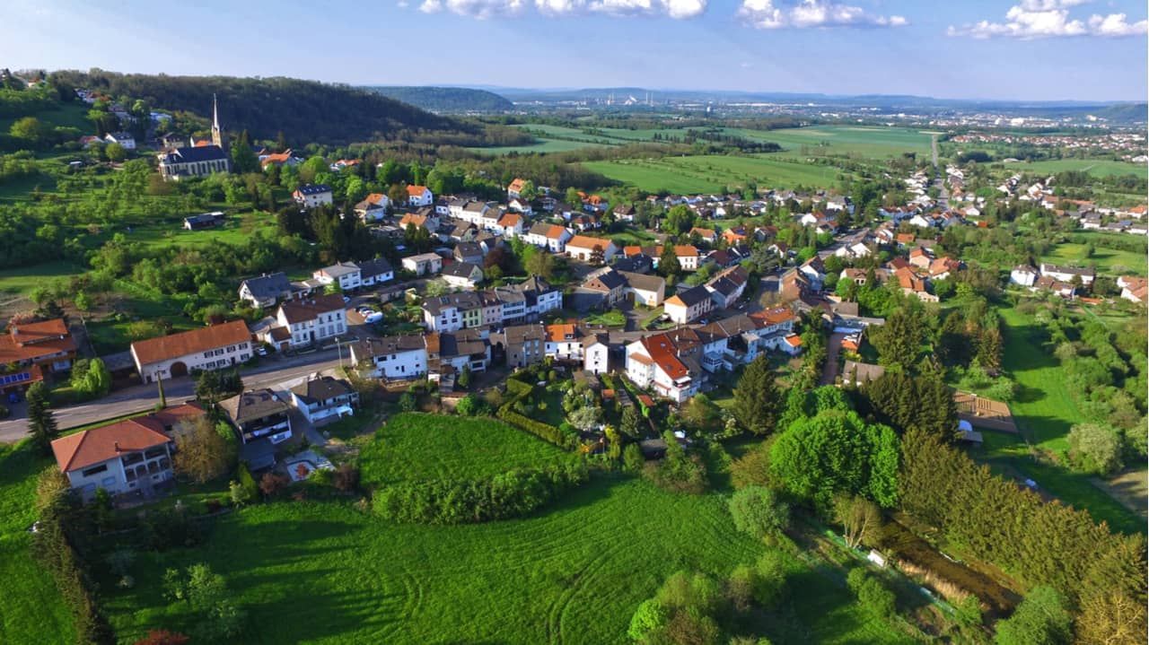 Aerial view of a European village with houses, green fields, and a church on a hillside under a sunny sky.