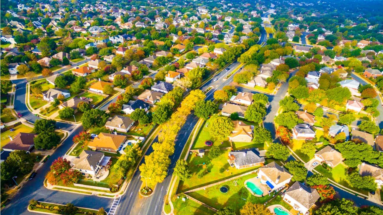 Aerial view of suburban houses with trees, roads, and a blue sky.