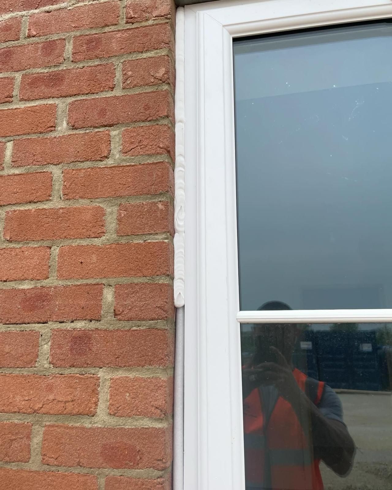 White window frame next to red brick wall with caulk along the side. A person in a safety vest is reflected in the window.