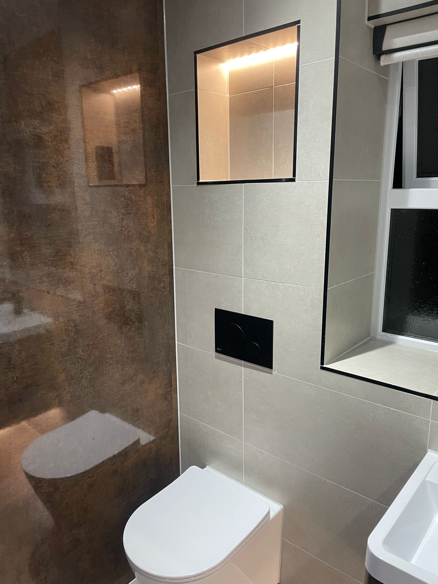 Modern bathroom with toilet, recessed shelf, and window, featuring neutral-toned tiles and black accents.