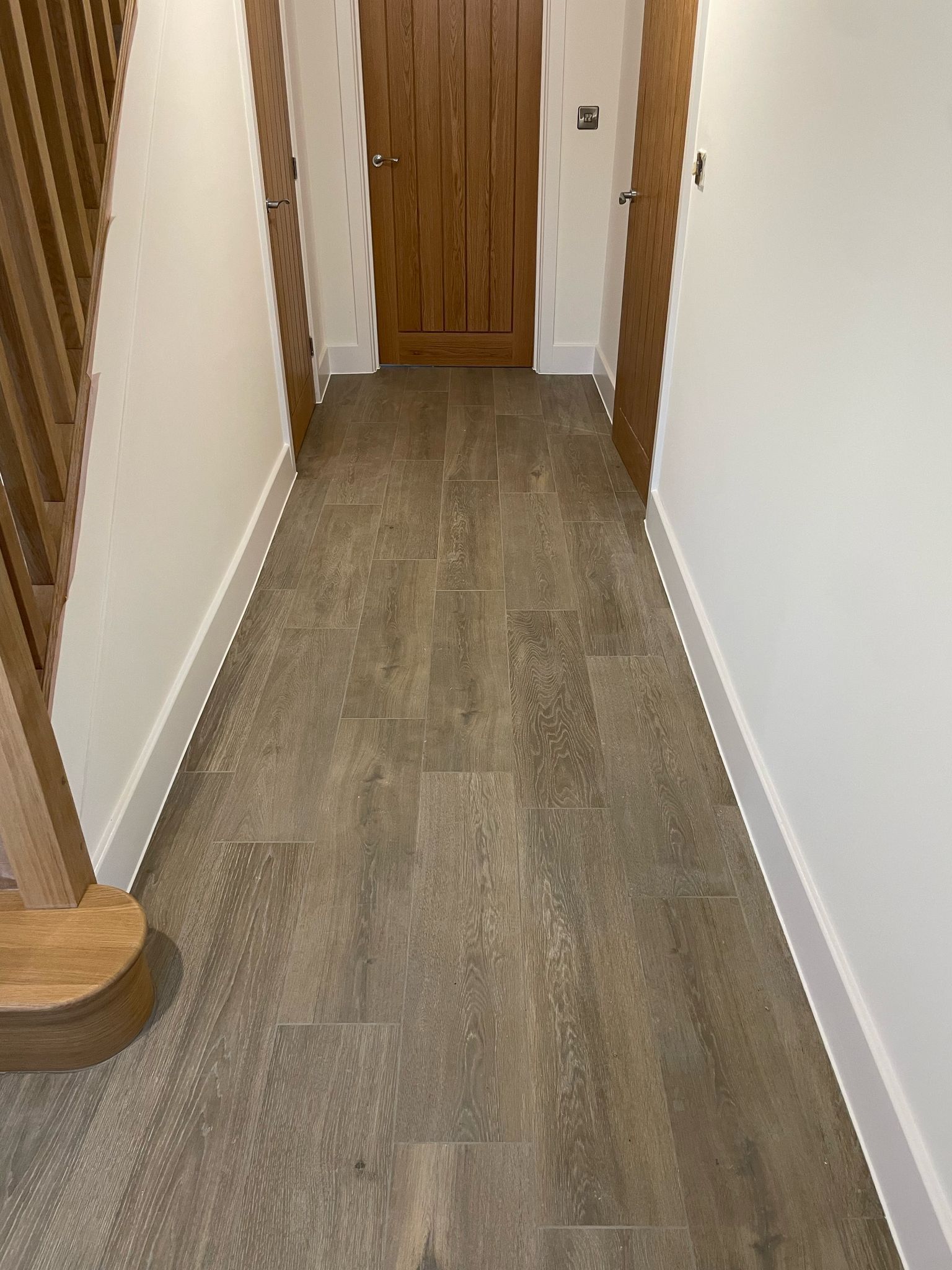 Narrow hallway with wood-look floor, white walls, wooden door at the end, and stairs on the left.
