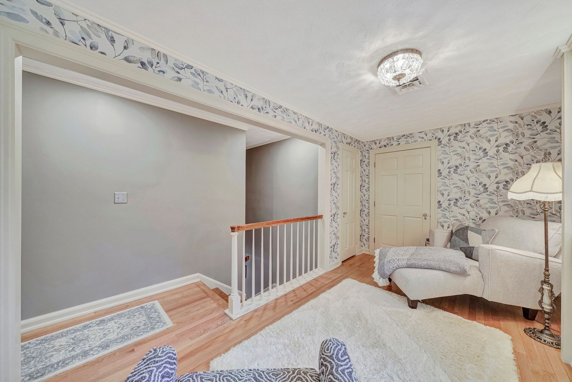 Cozy room with white armchair, patterned wallpaper, fluffy rug, hardwood floors, and a railing.