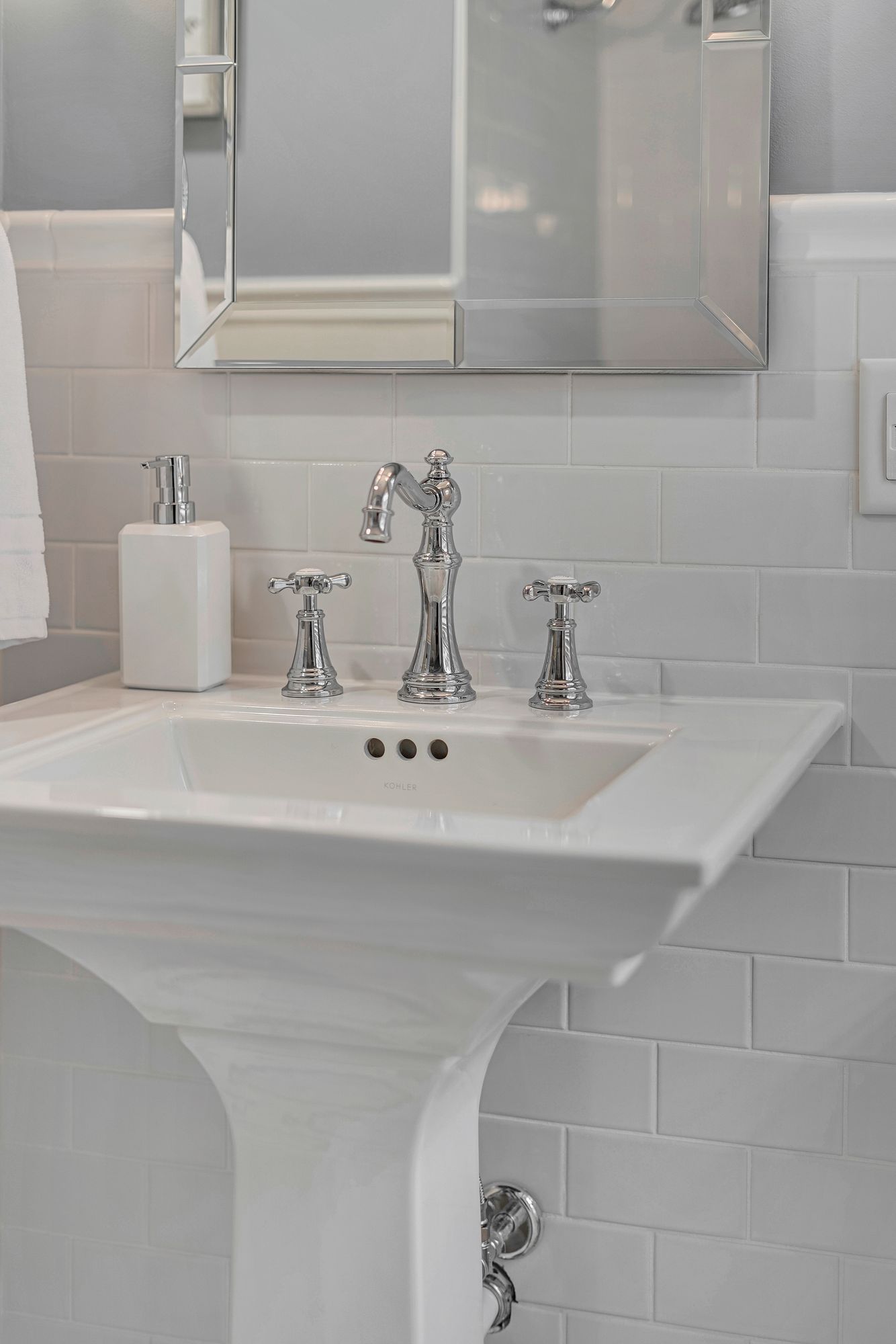 White pedestal sink with chrome faucet, soap dispenser, and mirror in a bathroom with white subway tile.