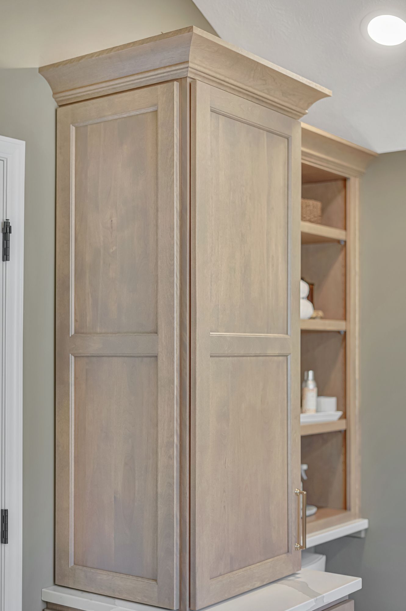 Tall, light-wood bathroom cabinet with closed doors and a shelf unit next to it, against a neutral wall.
