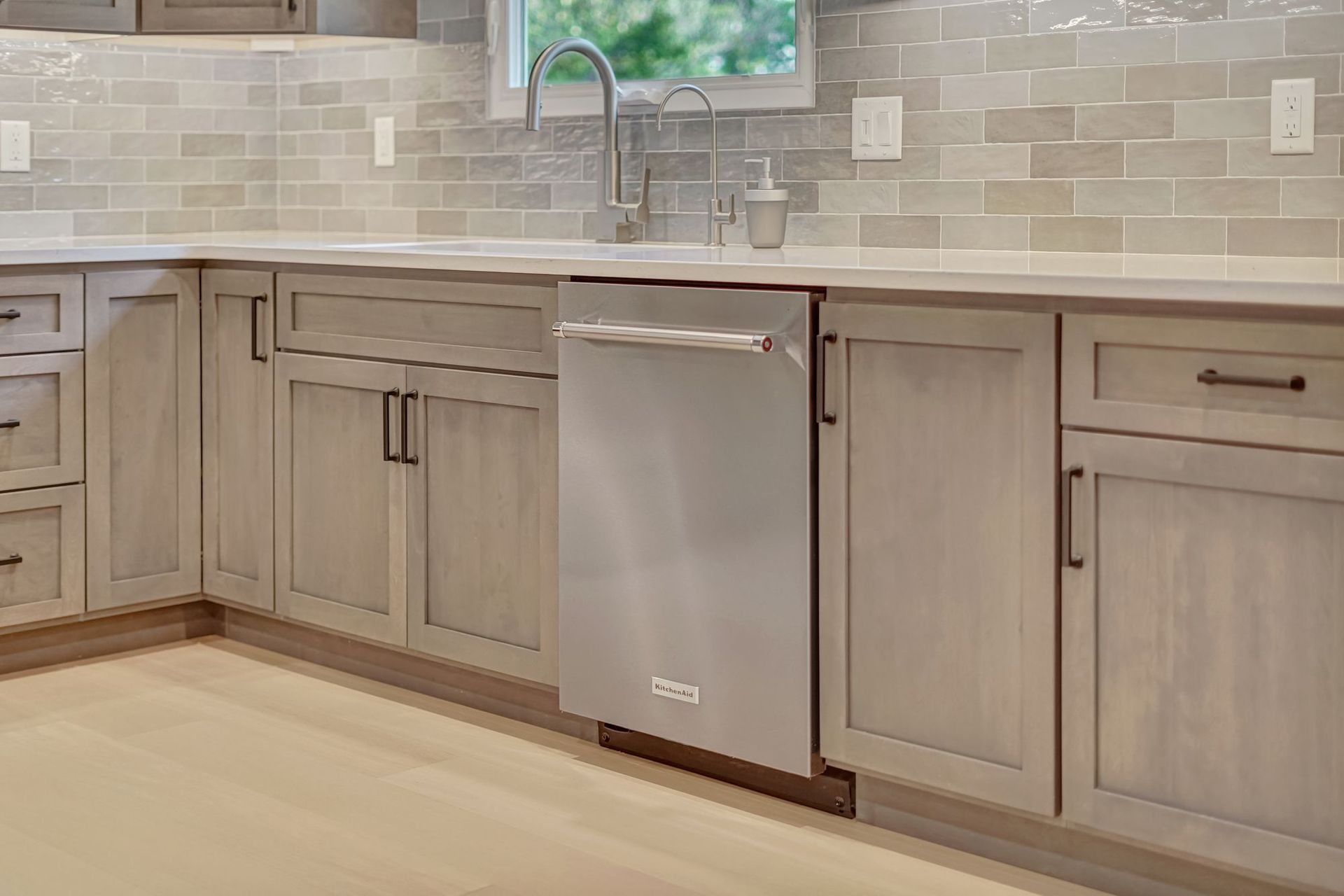 Kitchen with light wood cabinets, stainless steel dishwasher, light countertop, and tiled backsplash.