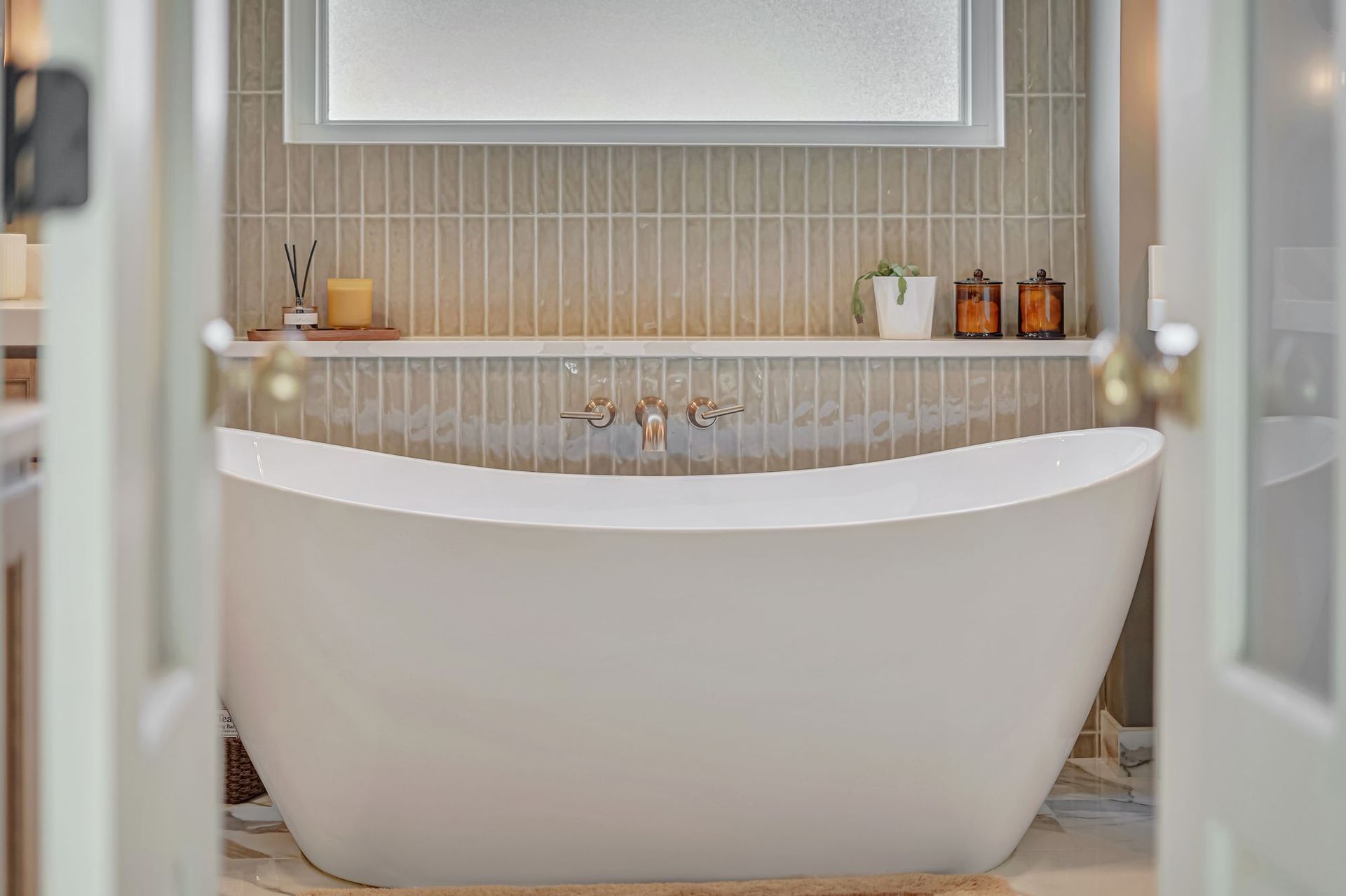 Freestanding white bathtub in a bathroom, framed by open doors; neutral tile backsplash and shelf.
