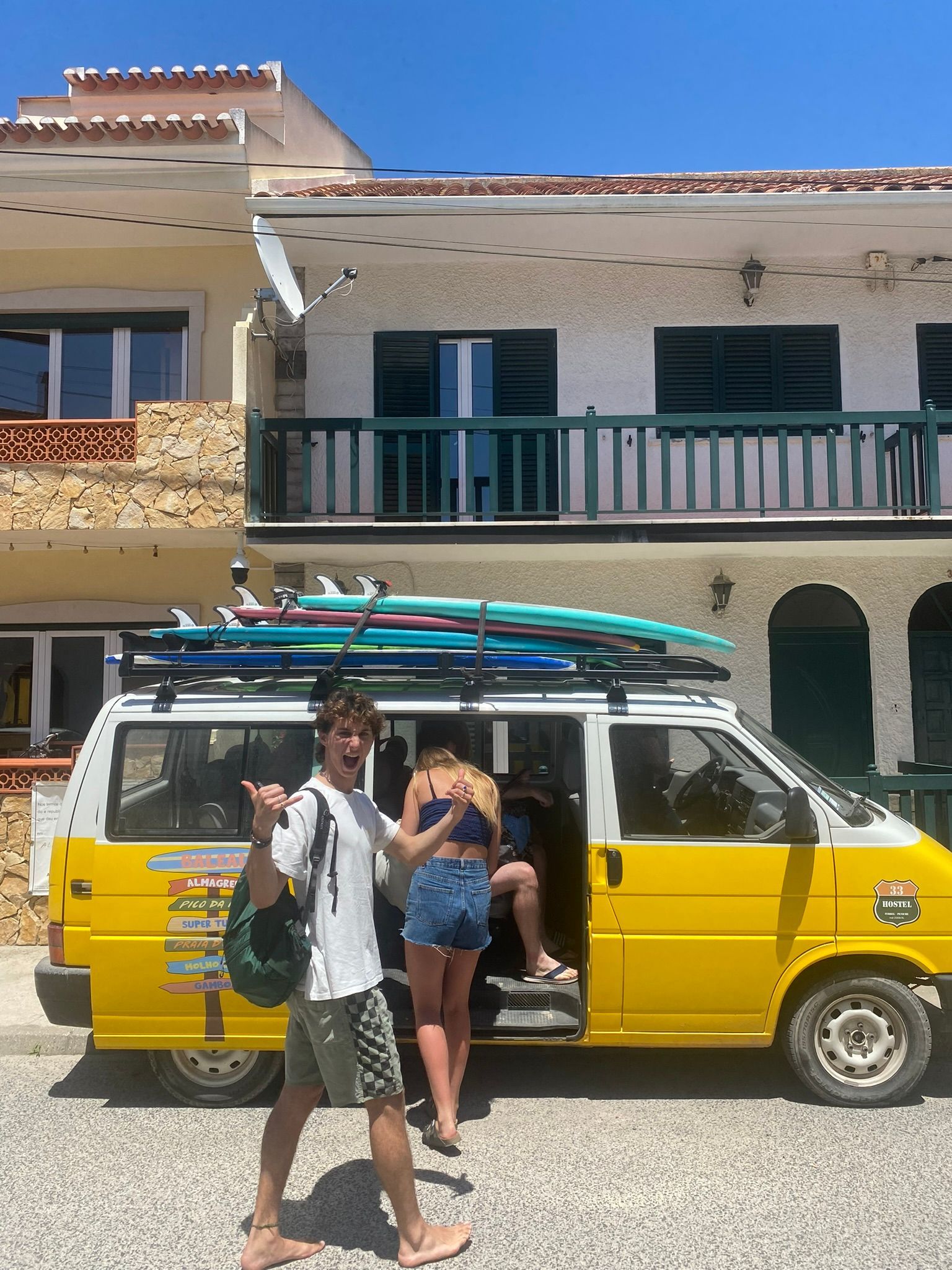 A man and a woman are standing in front of a yellow van with surfboards on the roof.