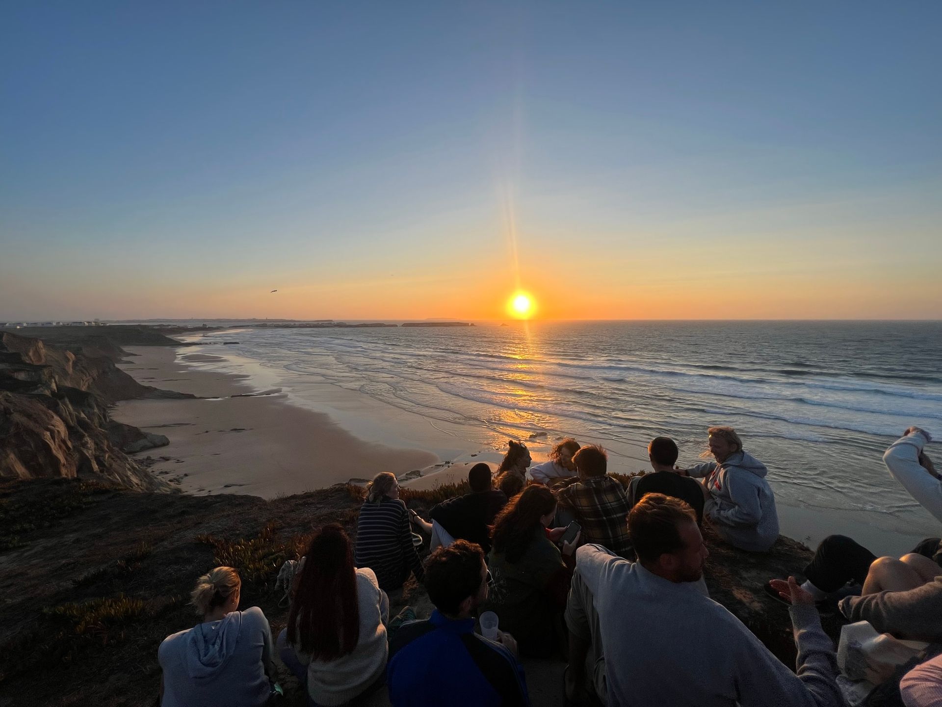 A group of people are sitting on top of a hill watching the sun set over the ocean.