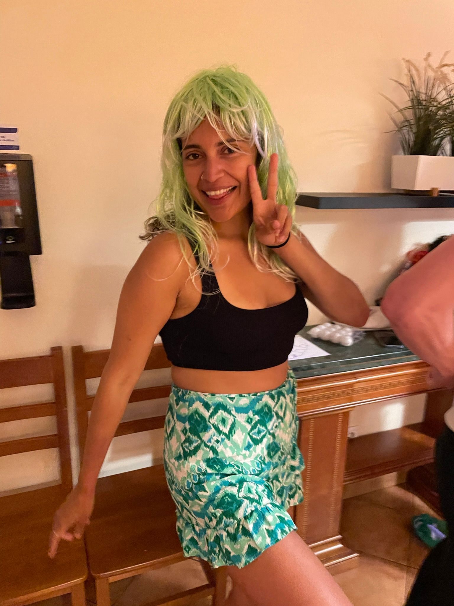 A woman wearing a green wig and a black top is giving a peace sign.