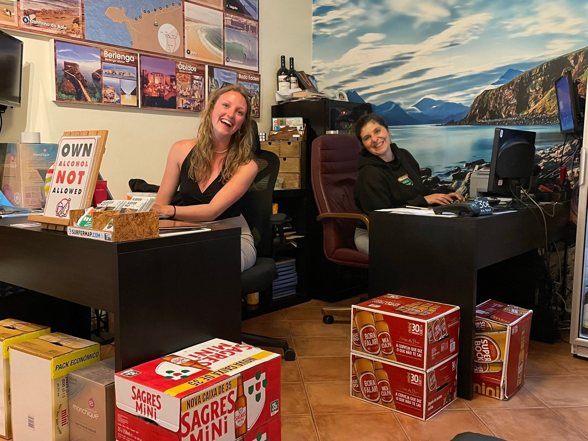 Two women are sitting at a desk in an office surrounded by boxes.