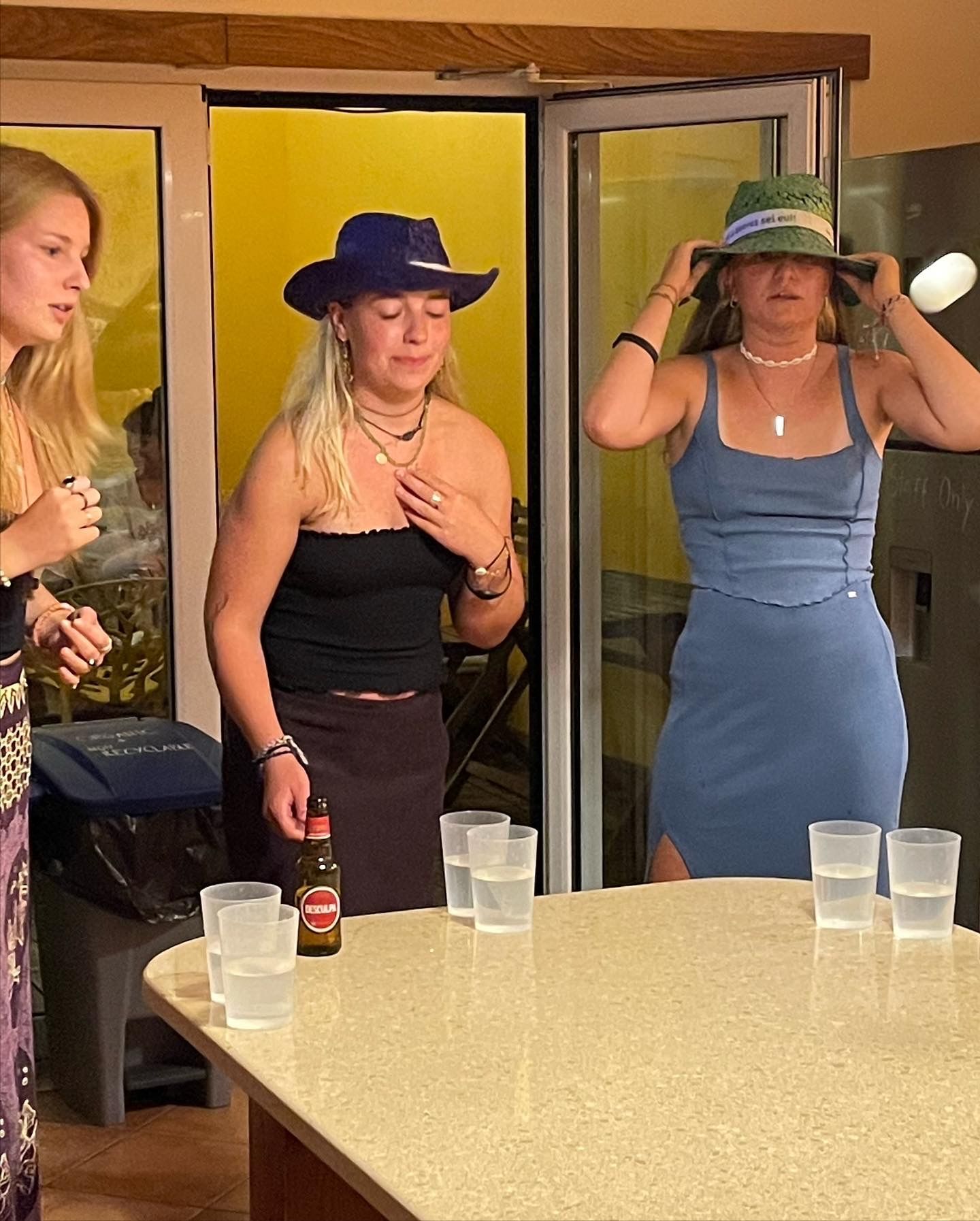 Three women are standing around a table with cups and a bottle of beer