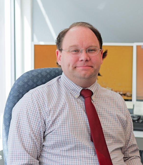Man wearing glasses and tie, sitting in office, smiling.