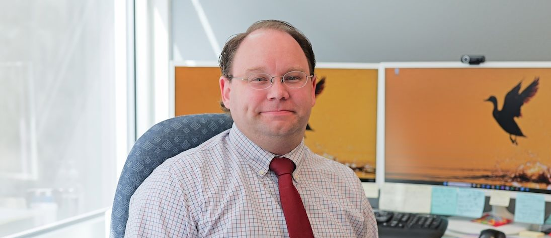 Man wearing glasses and a red tie, sitting at a desk with two computer monitors.