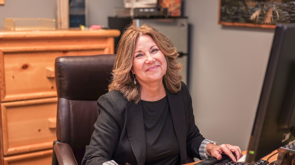 Woman smiling at a desk in an office setting, wearing a black blazer.