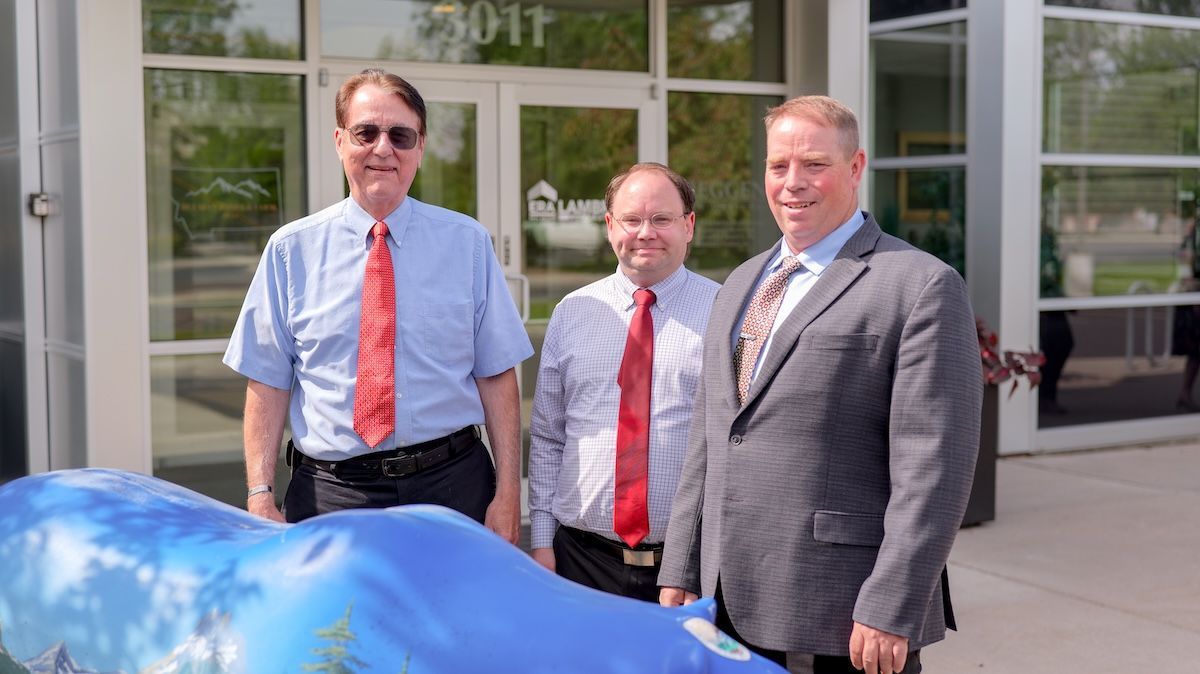 Three men in business attire pose outside a building, one with sunglasses, next to a blue art sculpture.