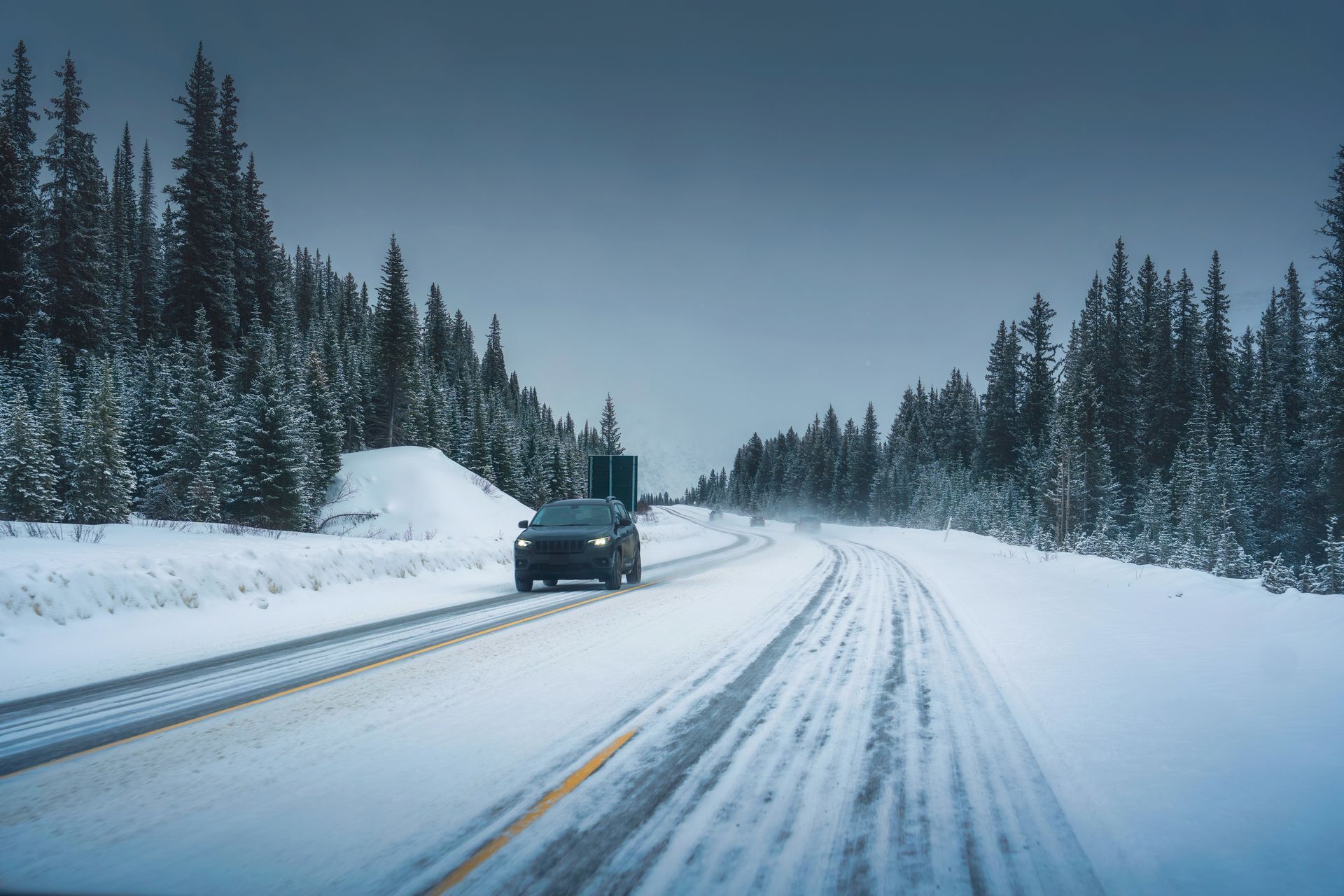 Black car driving on a snow-covered road through a forest with evergreens, under a gray sky.