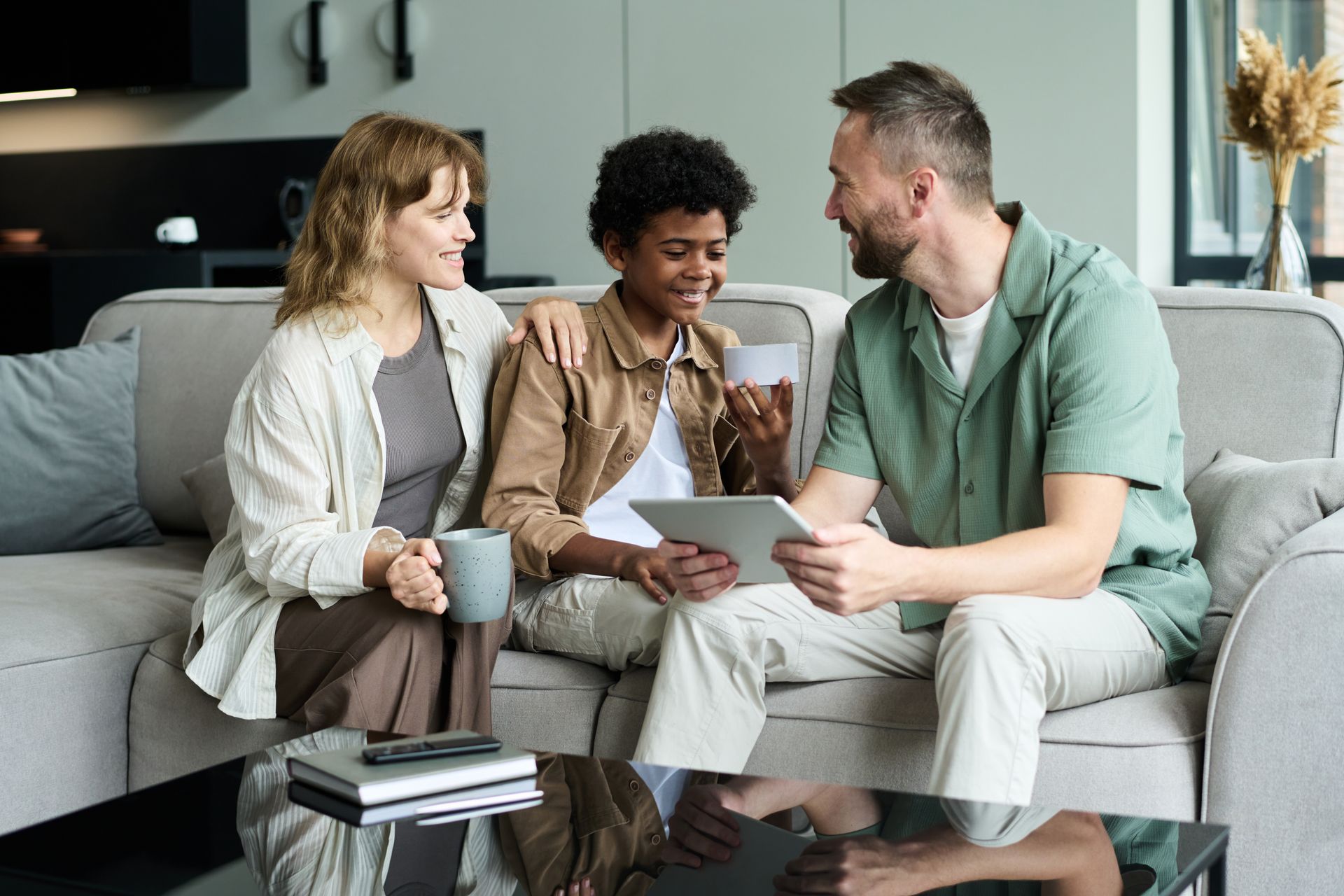 Family of three sitting on a couch, looking at a tablet, smiling.