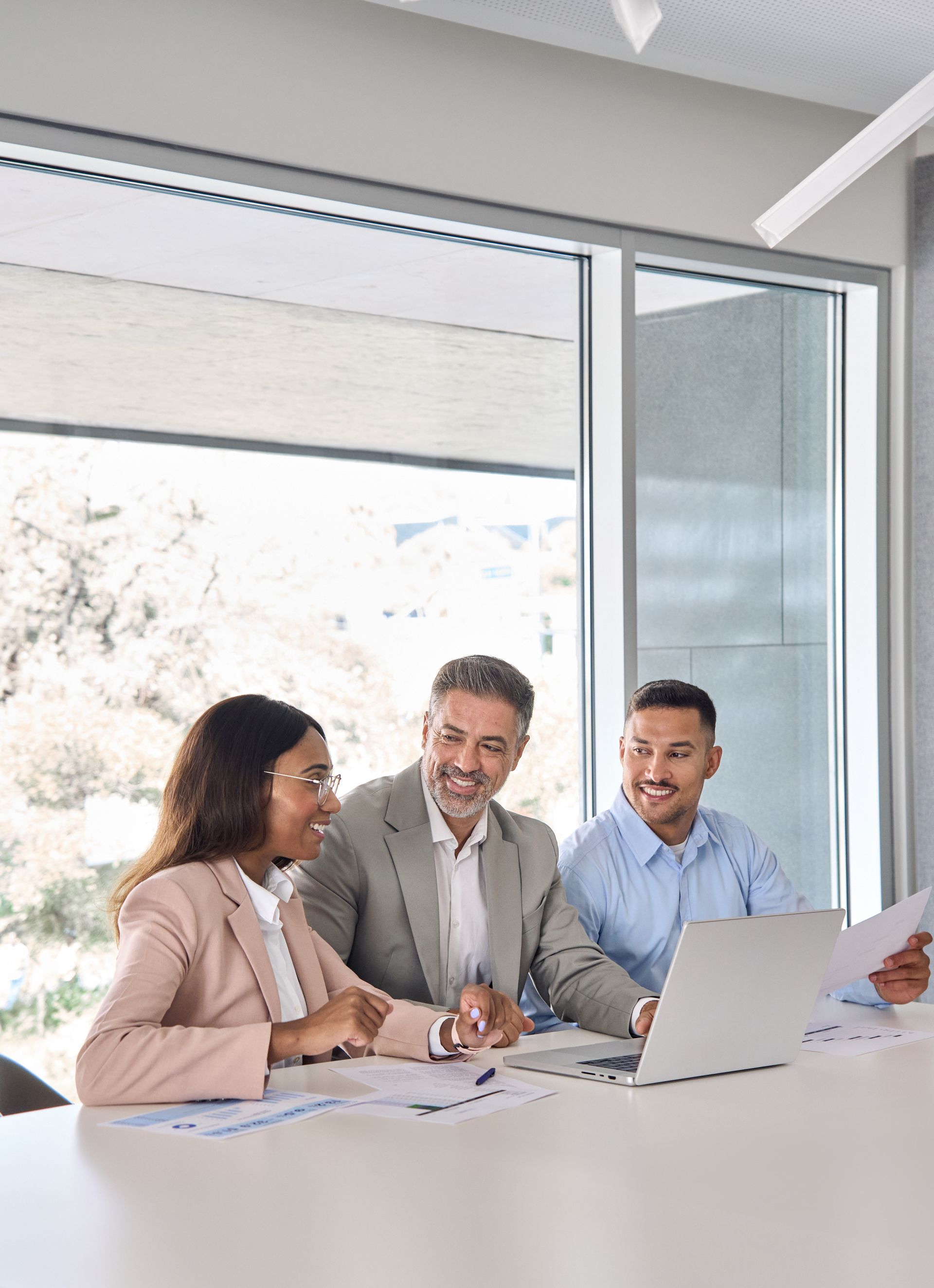 Three people reviewing documents and laptop at a table, smiling, in a modern office with large window.