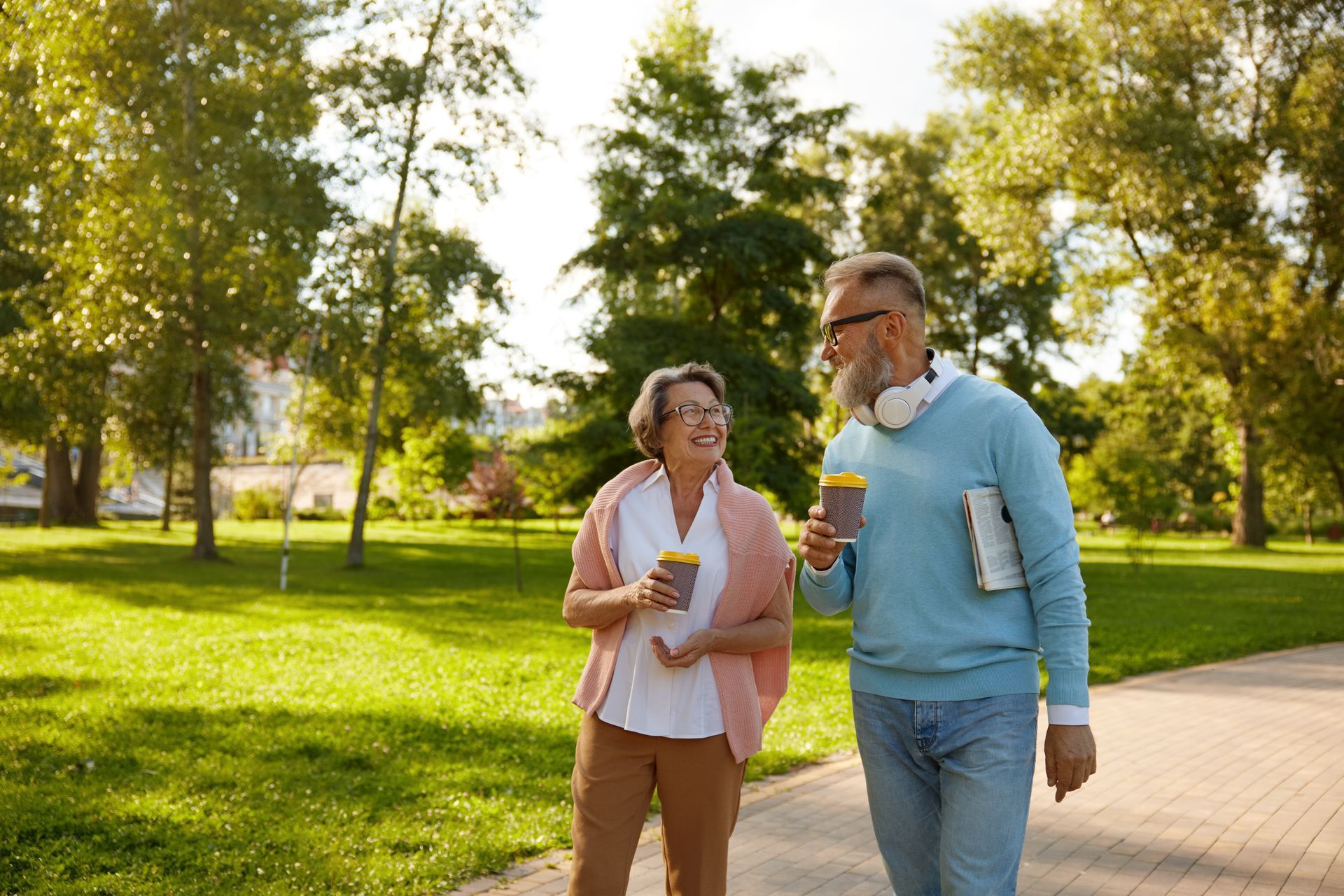 A couple walking and talking in a park, holding coffee cups. The man has a beard and headphones.