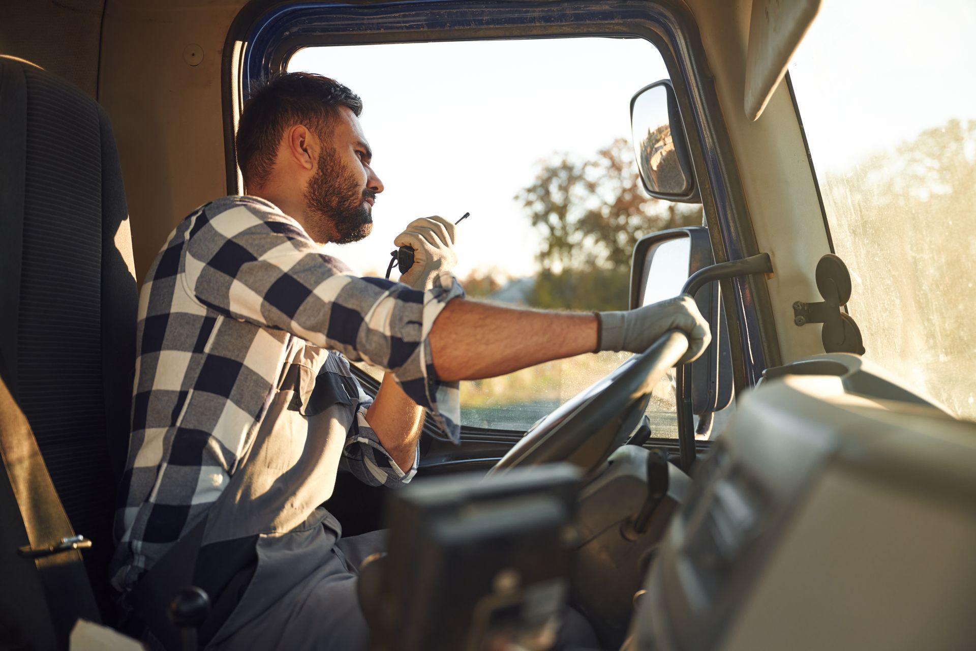 Truck driver in plaid shirt, holding a radio, driving, with sunlight streaming through the window.