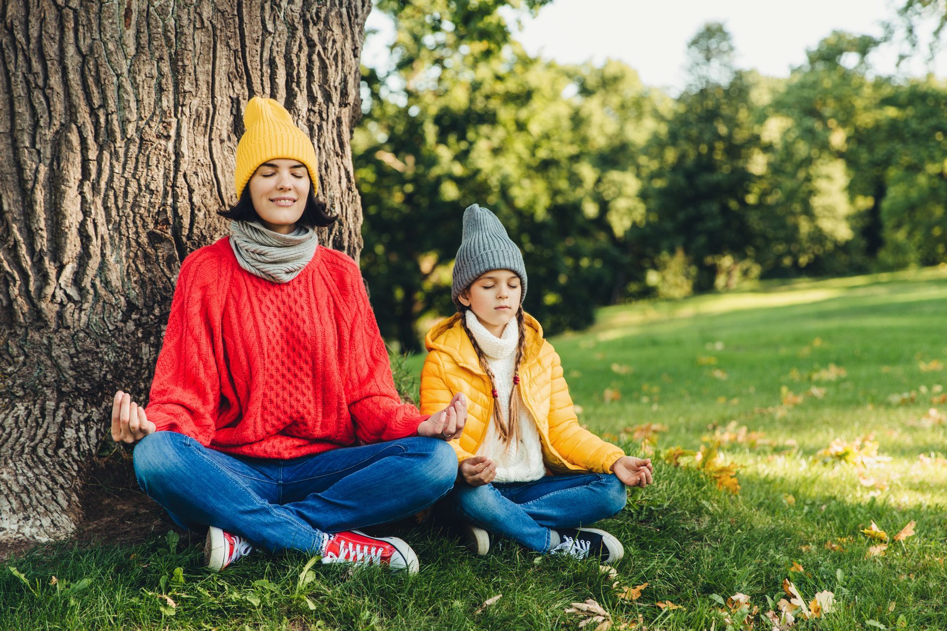 Woman and child meditating under a tree in a park; both wear hats, jeans, and colorful sweaters.