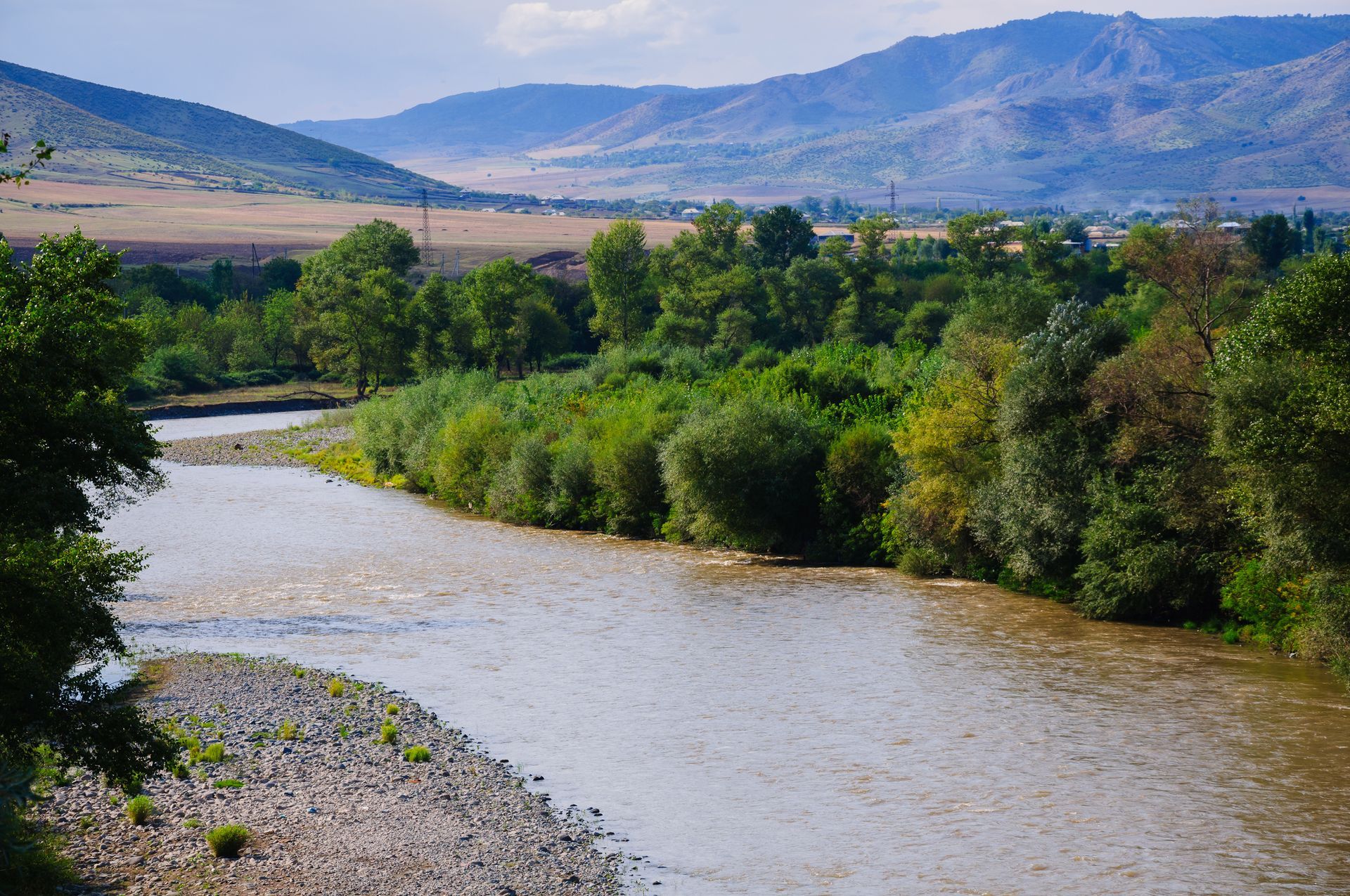 River flowing through lush green trees and rocky banks, mountains in the background.