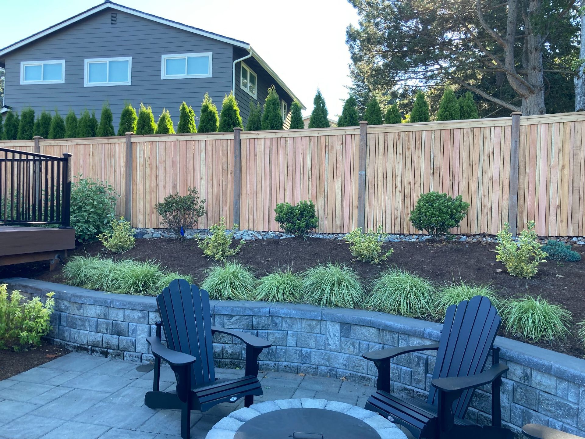Patio with Adirondack chairs, low stone wall, and wooden fence with landscaping. Patio with Adirondack chairs, low stone wall, and wooden fence with landscaping.