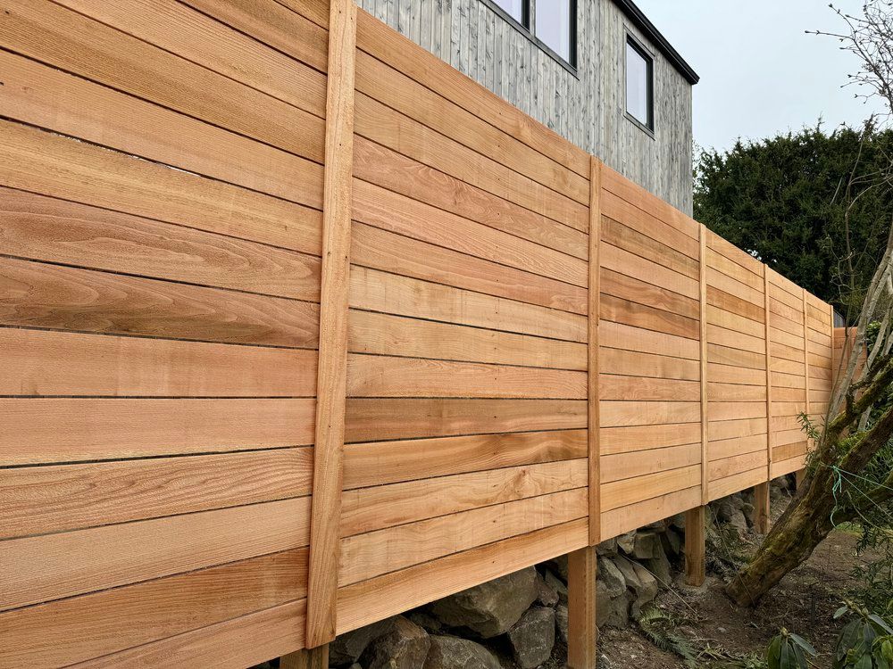 Wooden horizontal slat fence in front of a house, supported by posts on a rocky base.