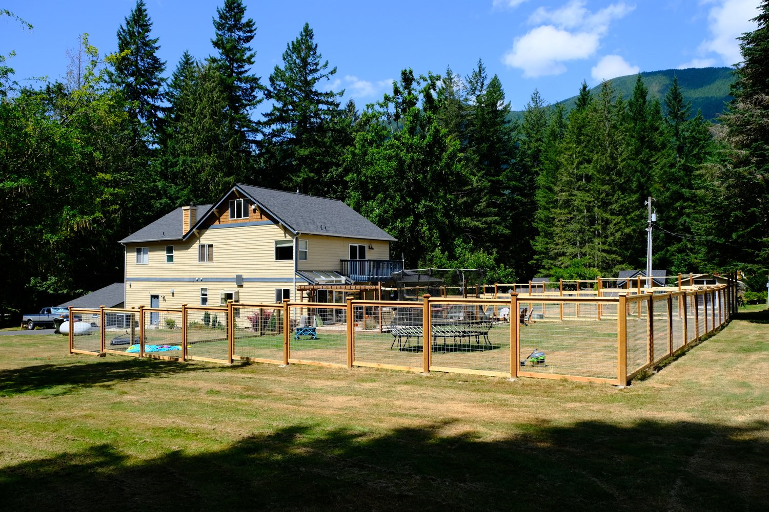 Yellow house with brown roof and wooden fence in a grassy yard surrounded by trees.