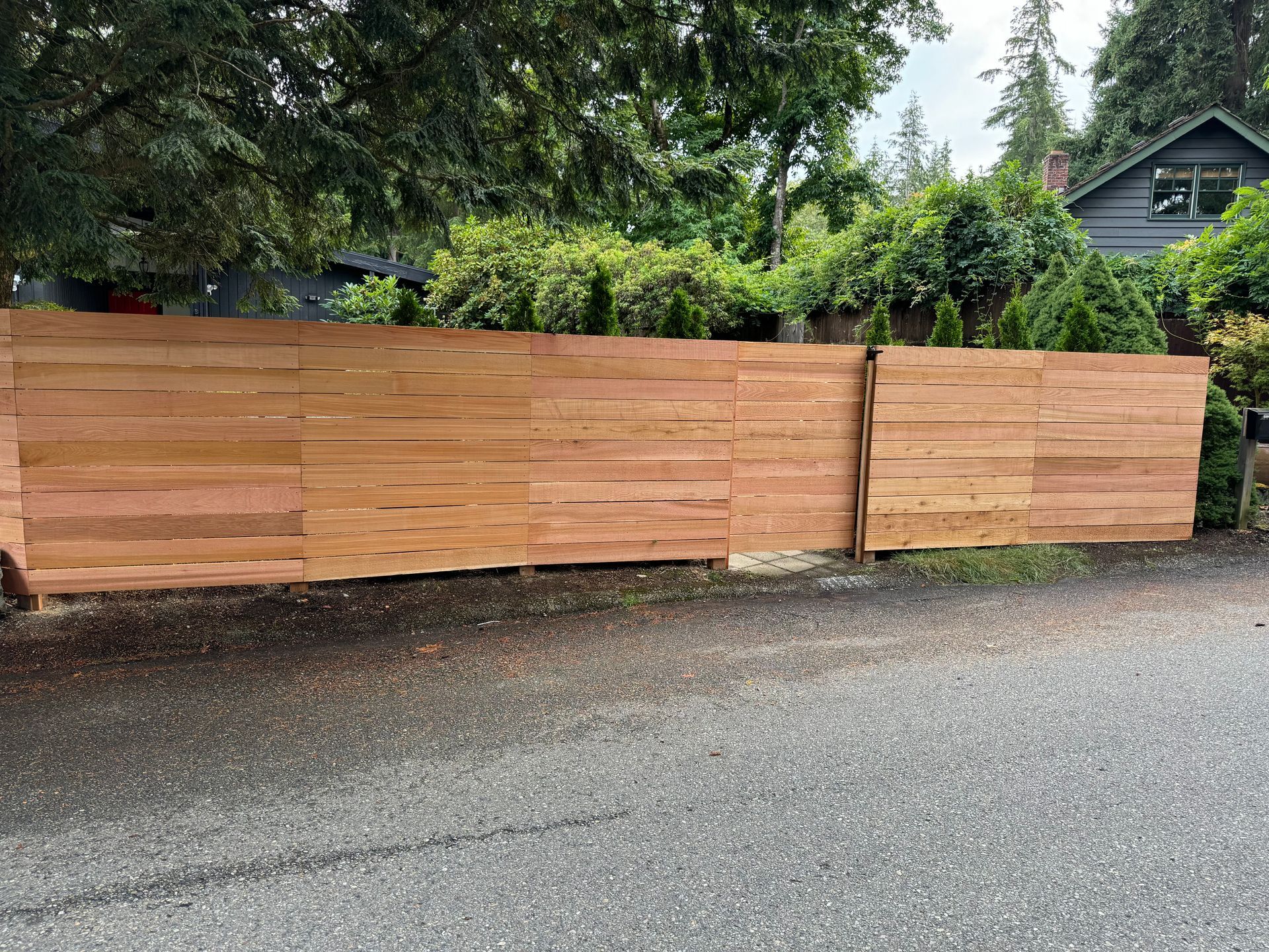 A wooden horizontal privacy fence spans across a residential area, with greenery and a house in the background.