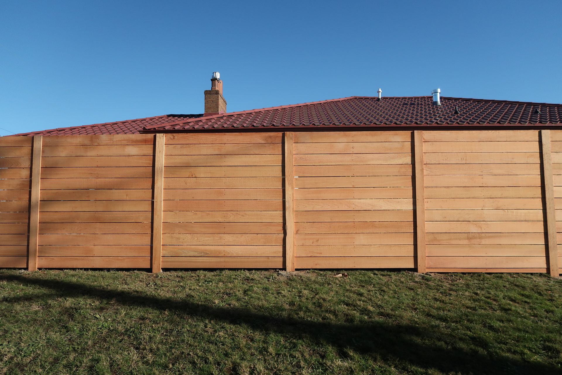 Wooden fence in front of a house with a red-tiled roof under a clear blue sky.