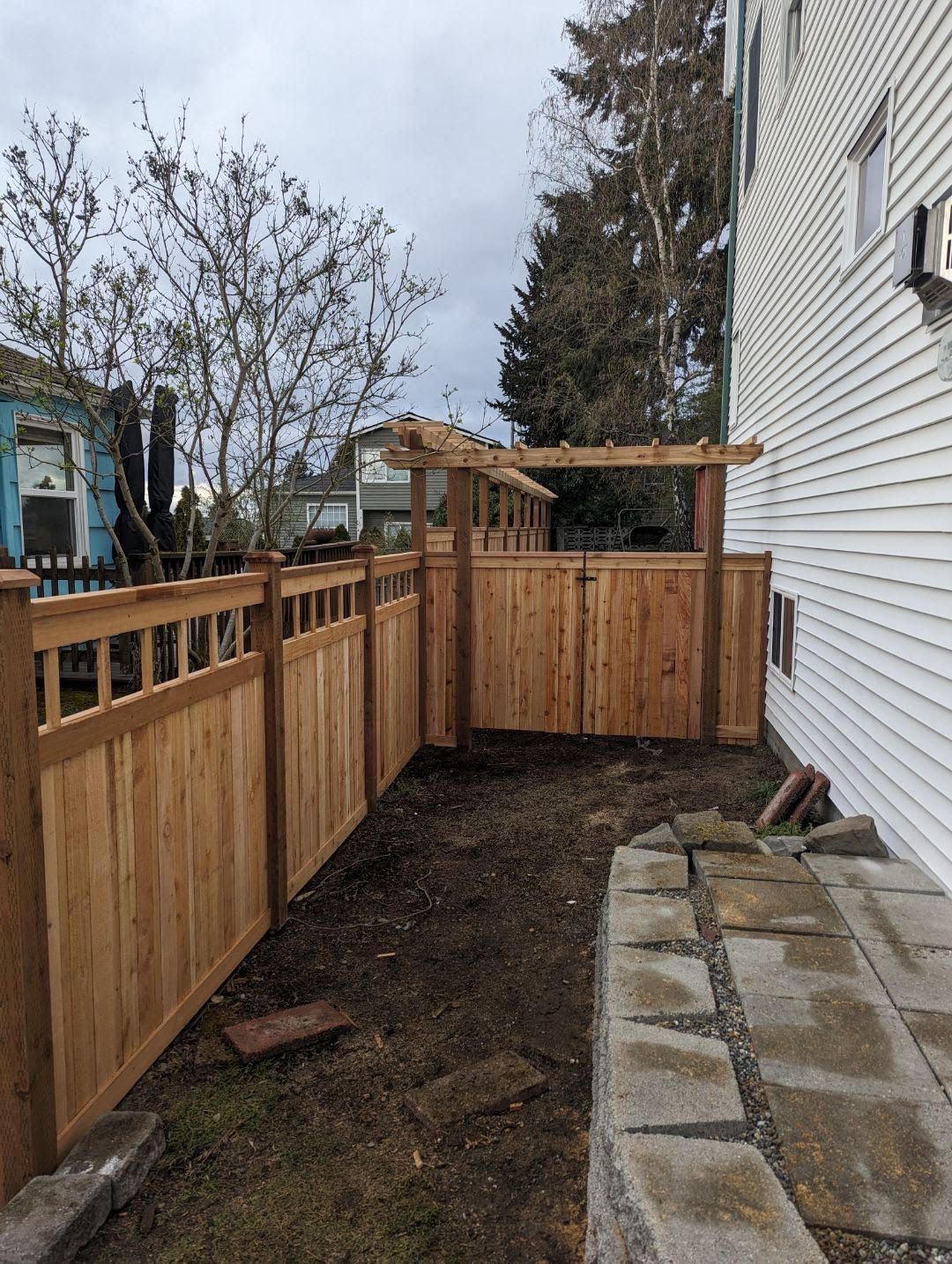 Wooden fence with gate and pergola next to a white house; yard with dirt and stone edging.
