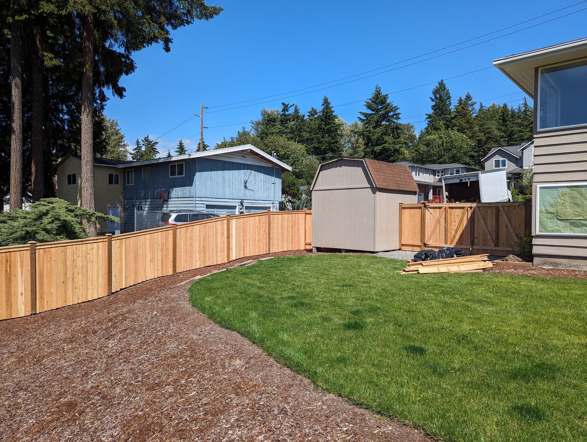 Wooden fence borders a lawn with a brown shed, blue house, and clear sky.