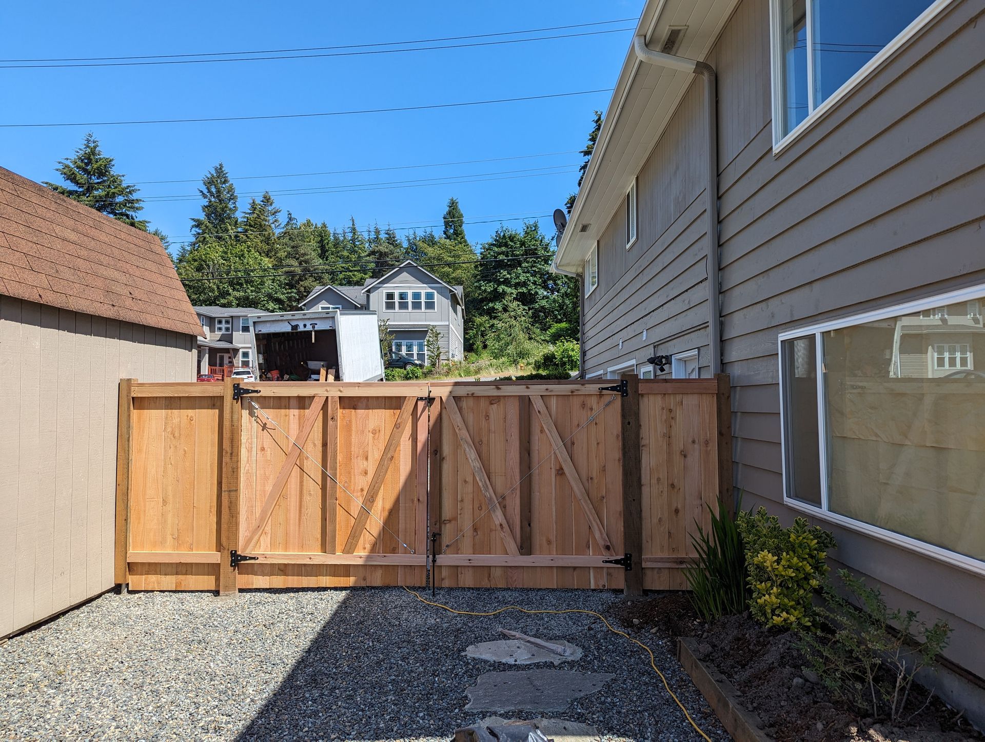 Wooden gate in a gravel yard, next to a beige shed and house with blue sky.