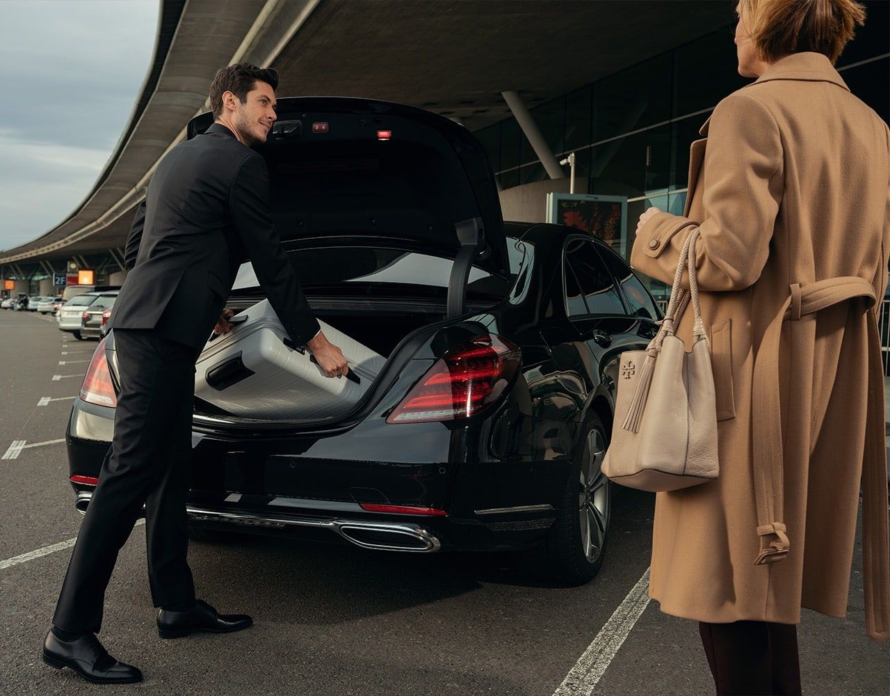 A man in a suit loads luggage into a black car's trunk for a woman at an airport.