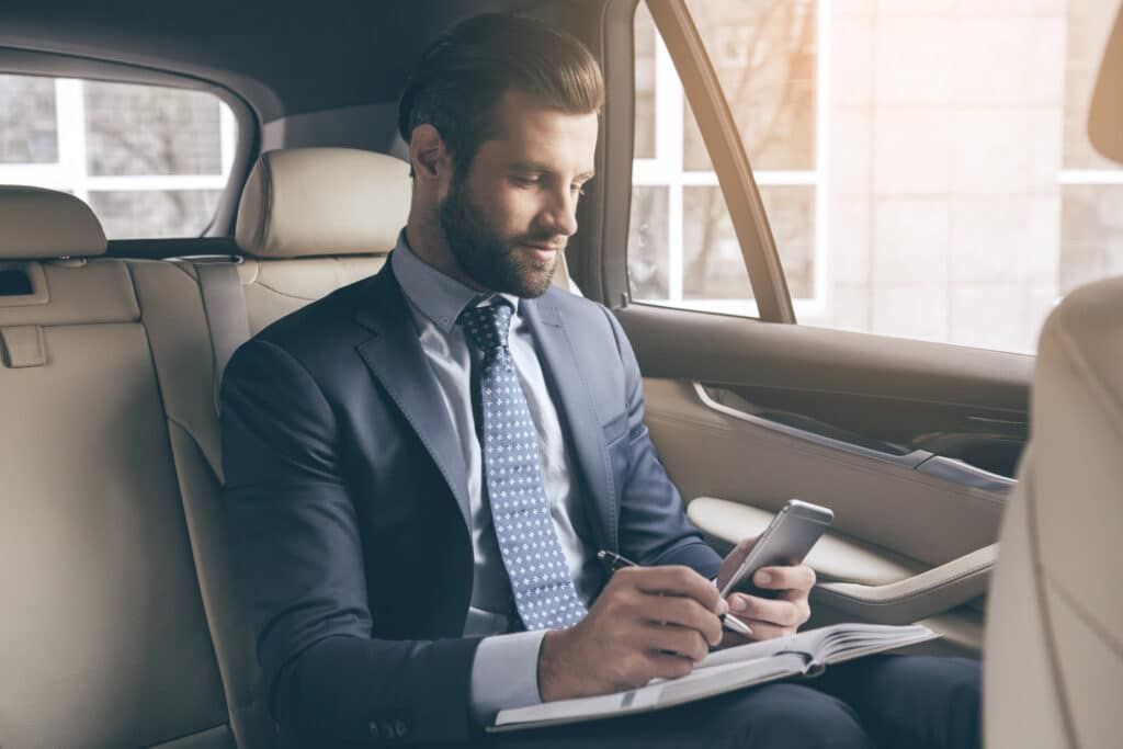 Man in suit, writing in notebook while using smartphone in car.